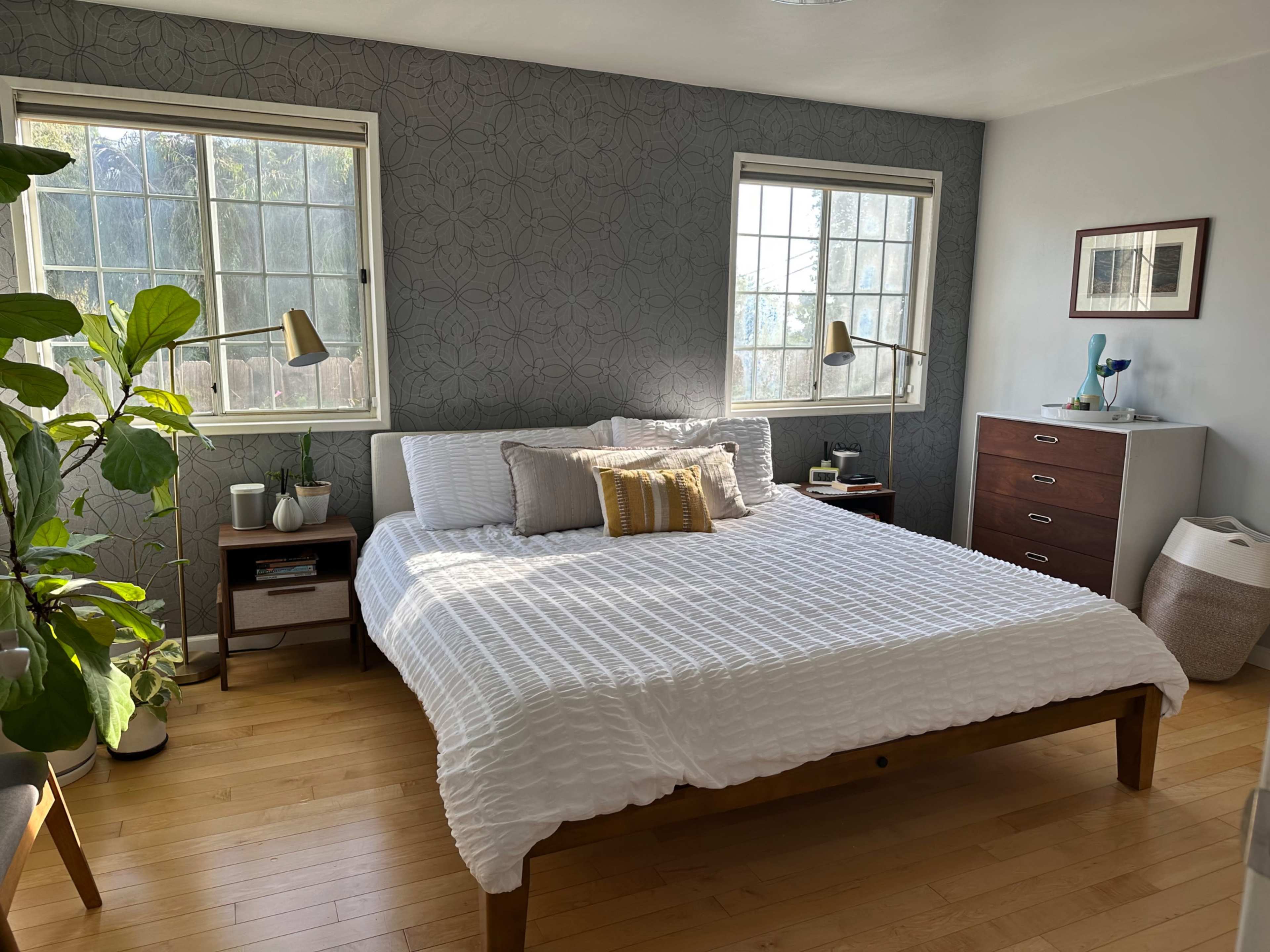 The image shows a well-lit bedroom featuring a bed with white bedding, two windows, a dresser, and a potted plant.