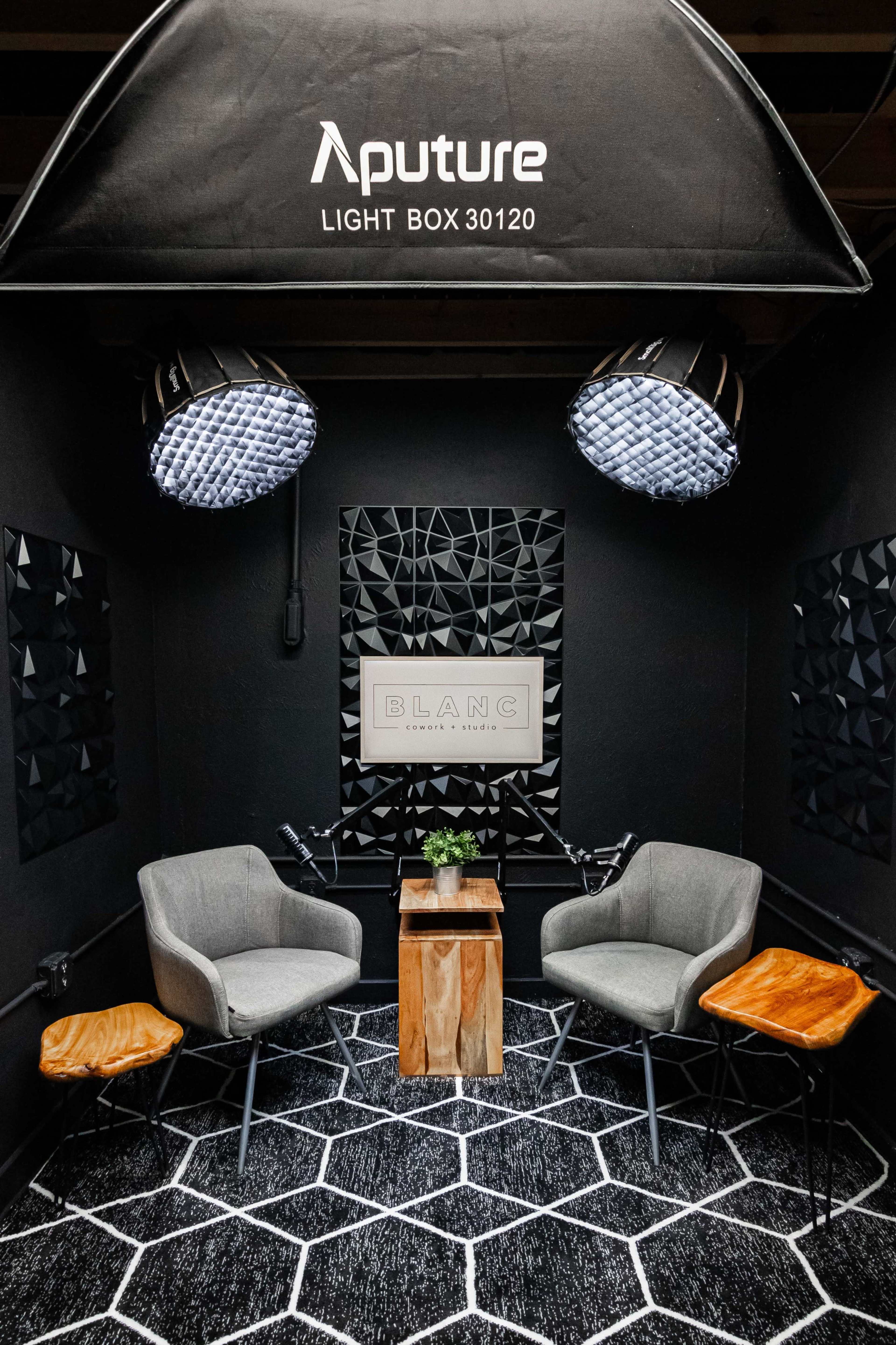 A photography setup features two gray chairs and a wooden table under a large lightbox, with geometric black wall panels in a dimly lit environment.