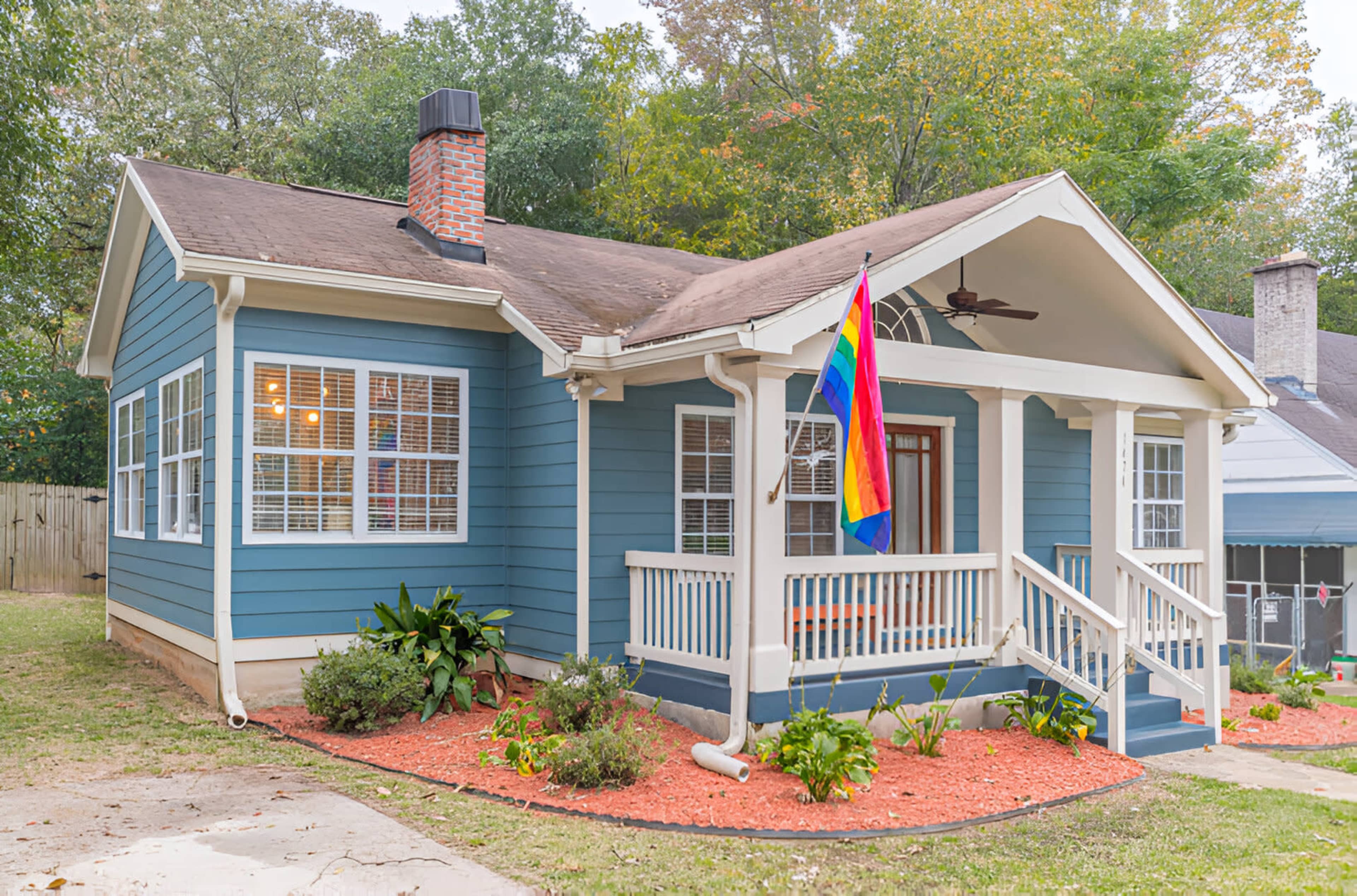 The image depicts a blue house with a front porch, a rainbow flag hanging from the porch, and landscaped flower beds in the yard.