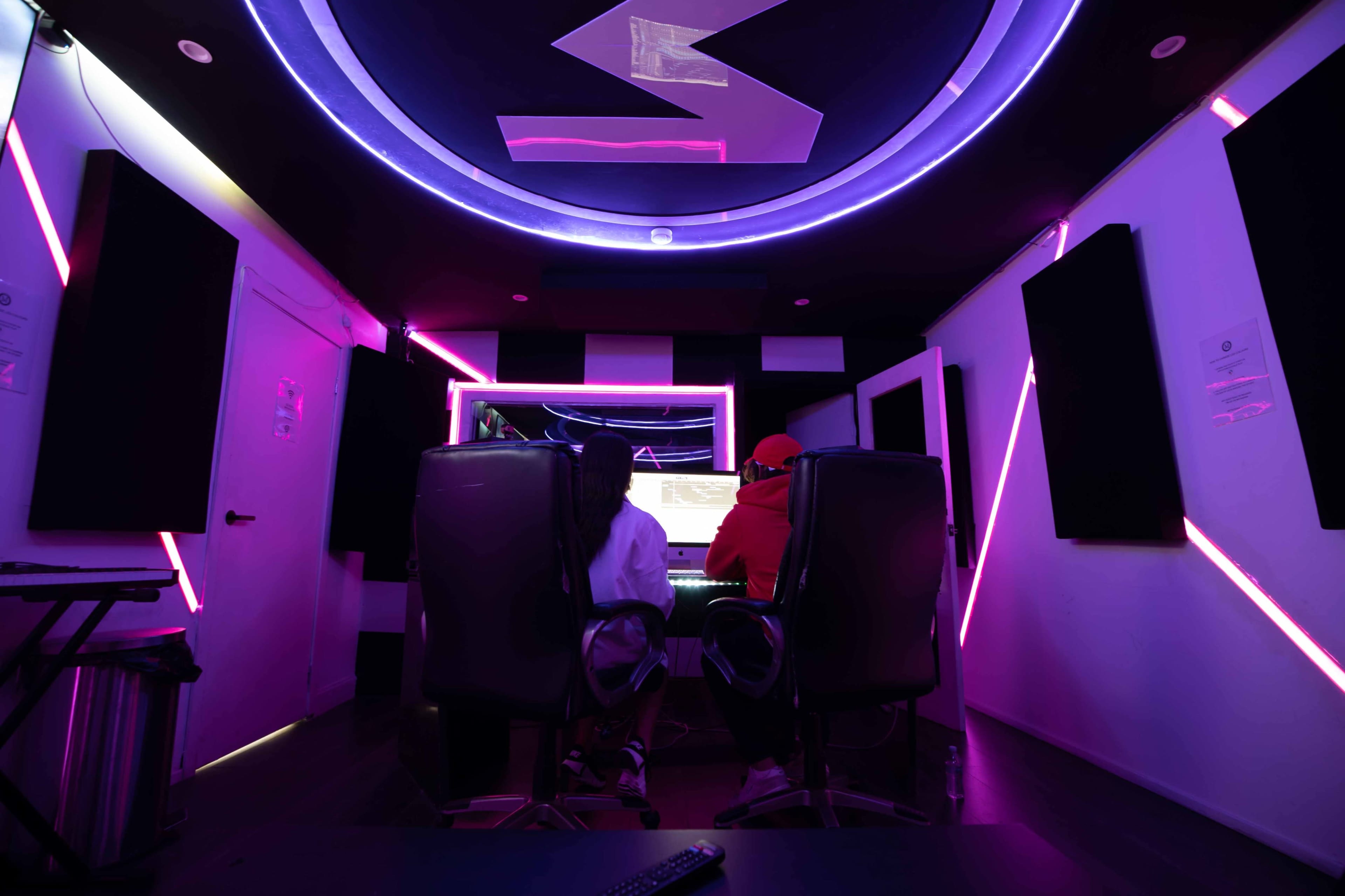 Two people sit in black chairs facing a brightly lit music production studio filled with neon accents.