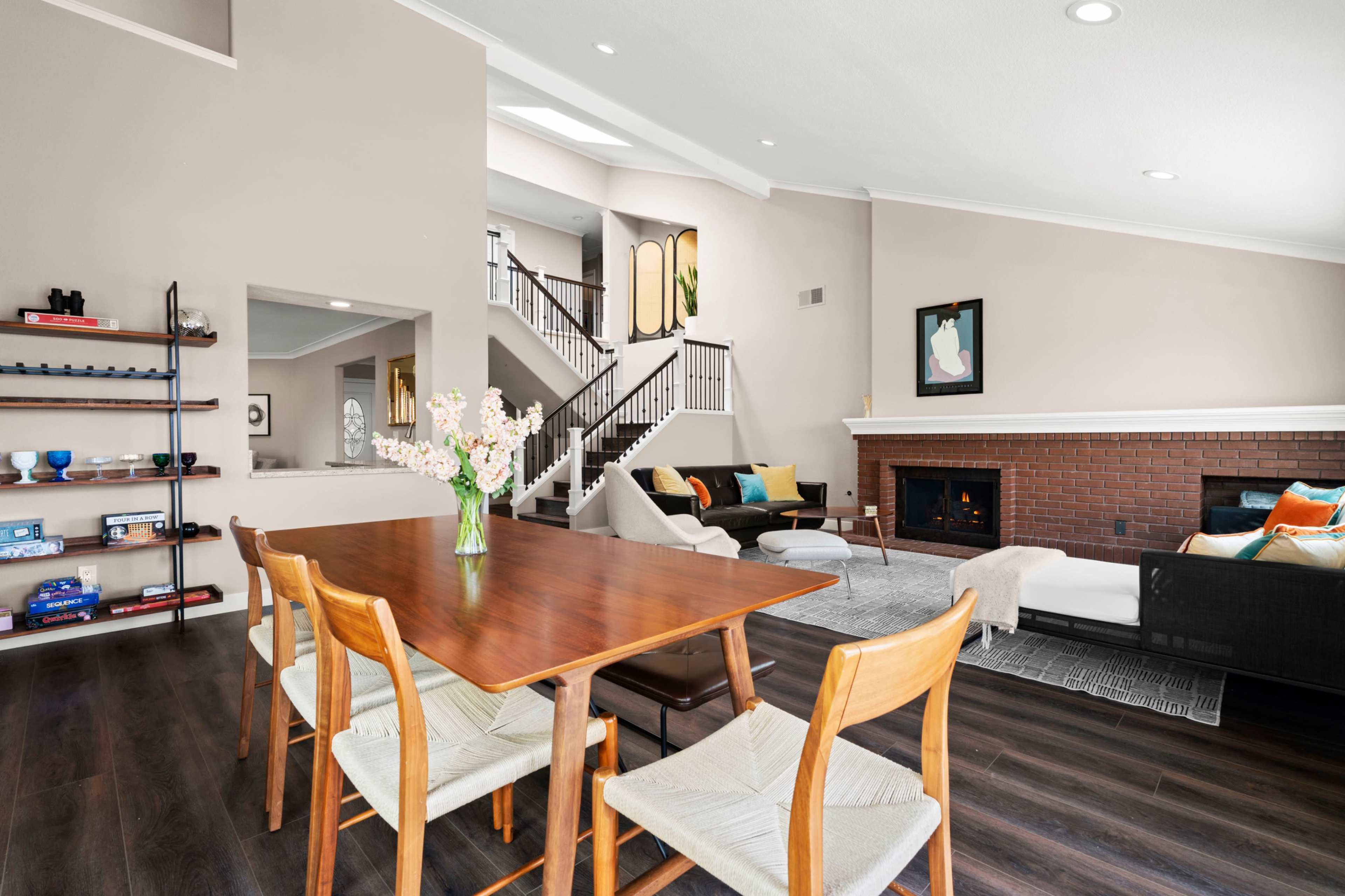 A modern living room features a dining table with chairs, a fireplace, and a staircase, with light-colored walls and wooden flooring.