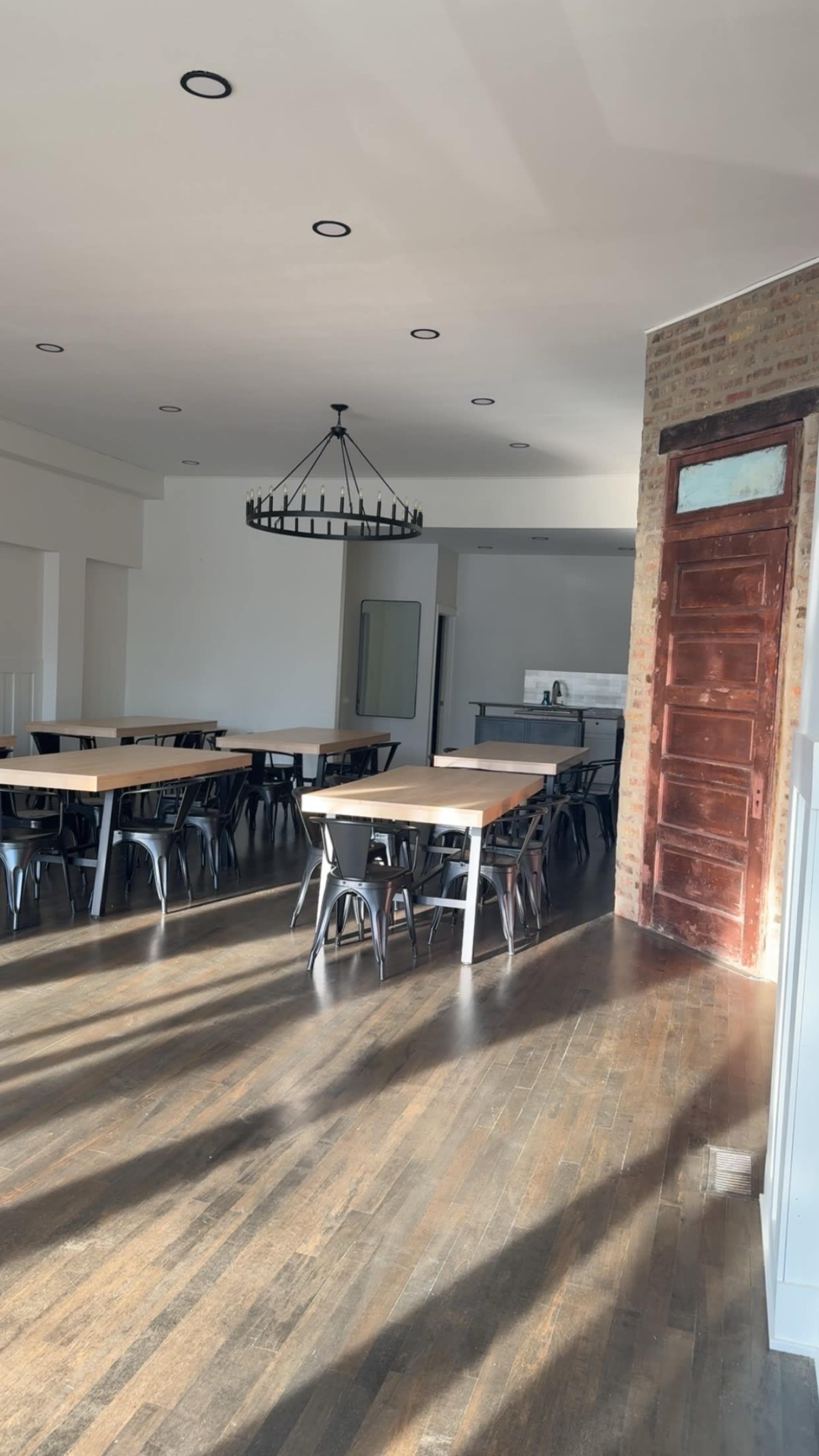 The image shows a spacious, well-lit interior with wooden floors, several long tables, and a large chandelier.