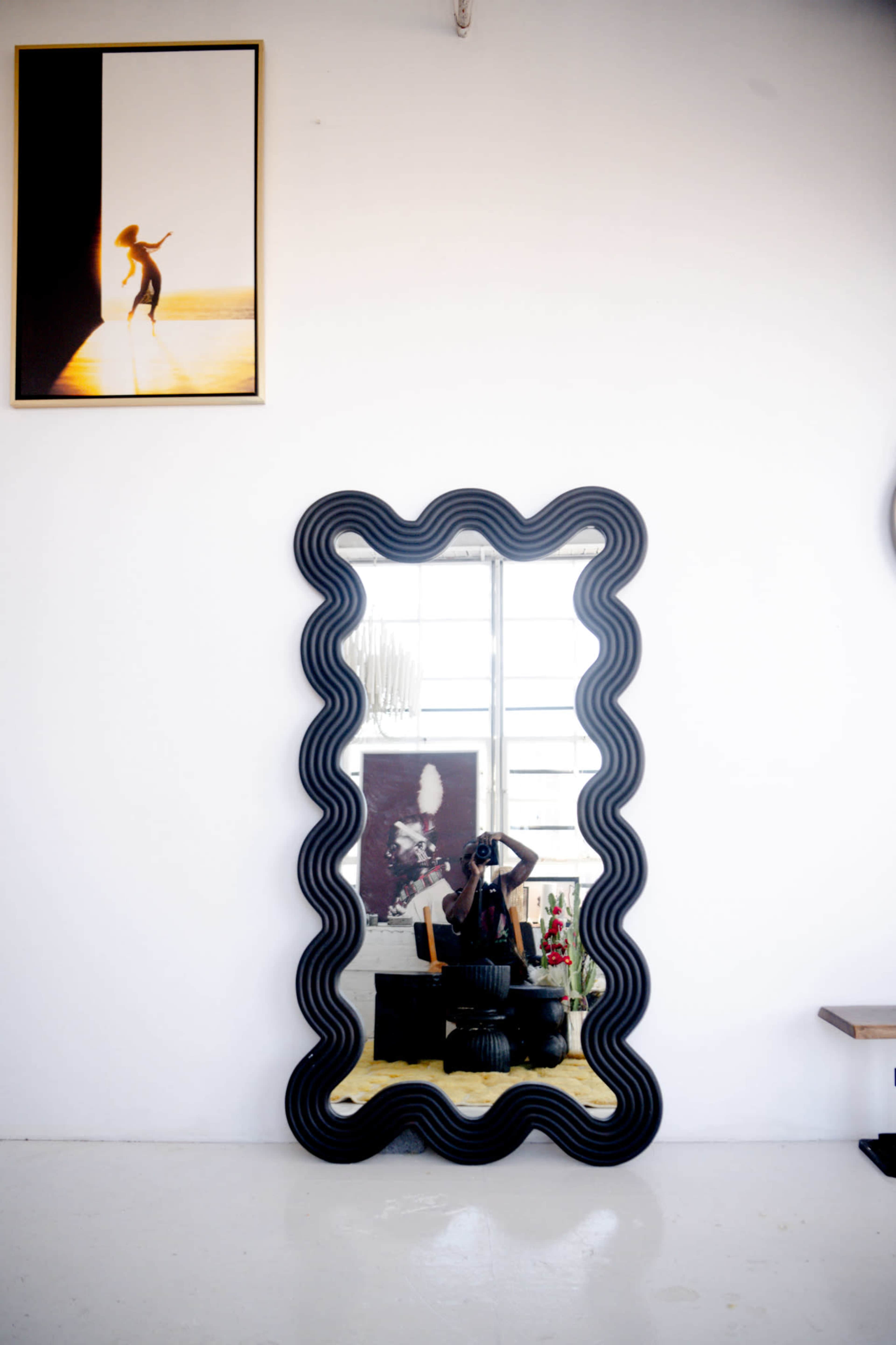 A large, black, wavy-framed mirror hanging on a white wall, reflecting a person taking a photo, alongside a framed picture of a figure in a sunset.