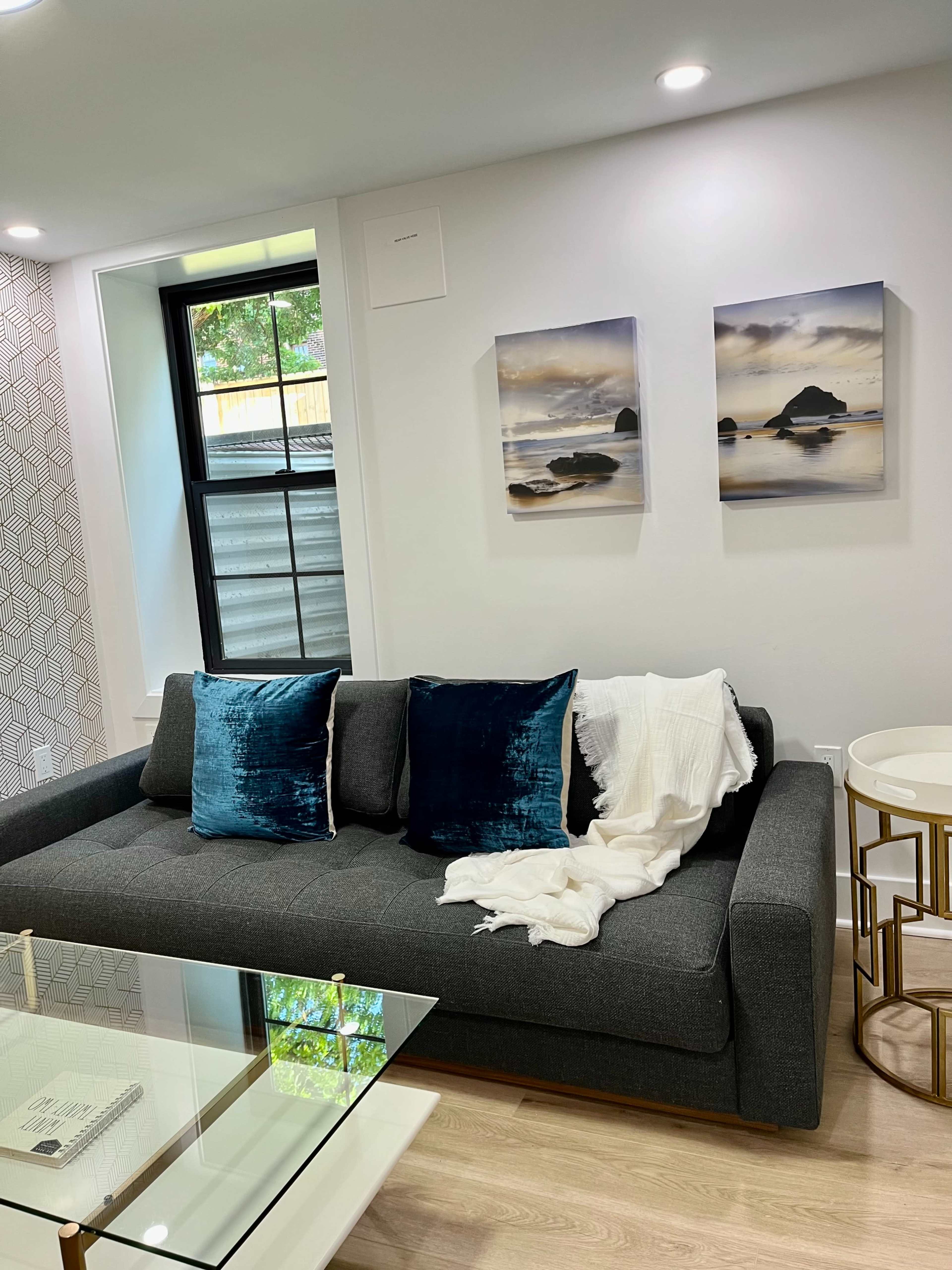 A gray sofa with two teal pillows and a white throw blanket is positioned in front of a glass coffee table, next to a window and two framed landscape paintings on the wall.
