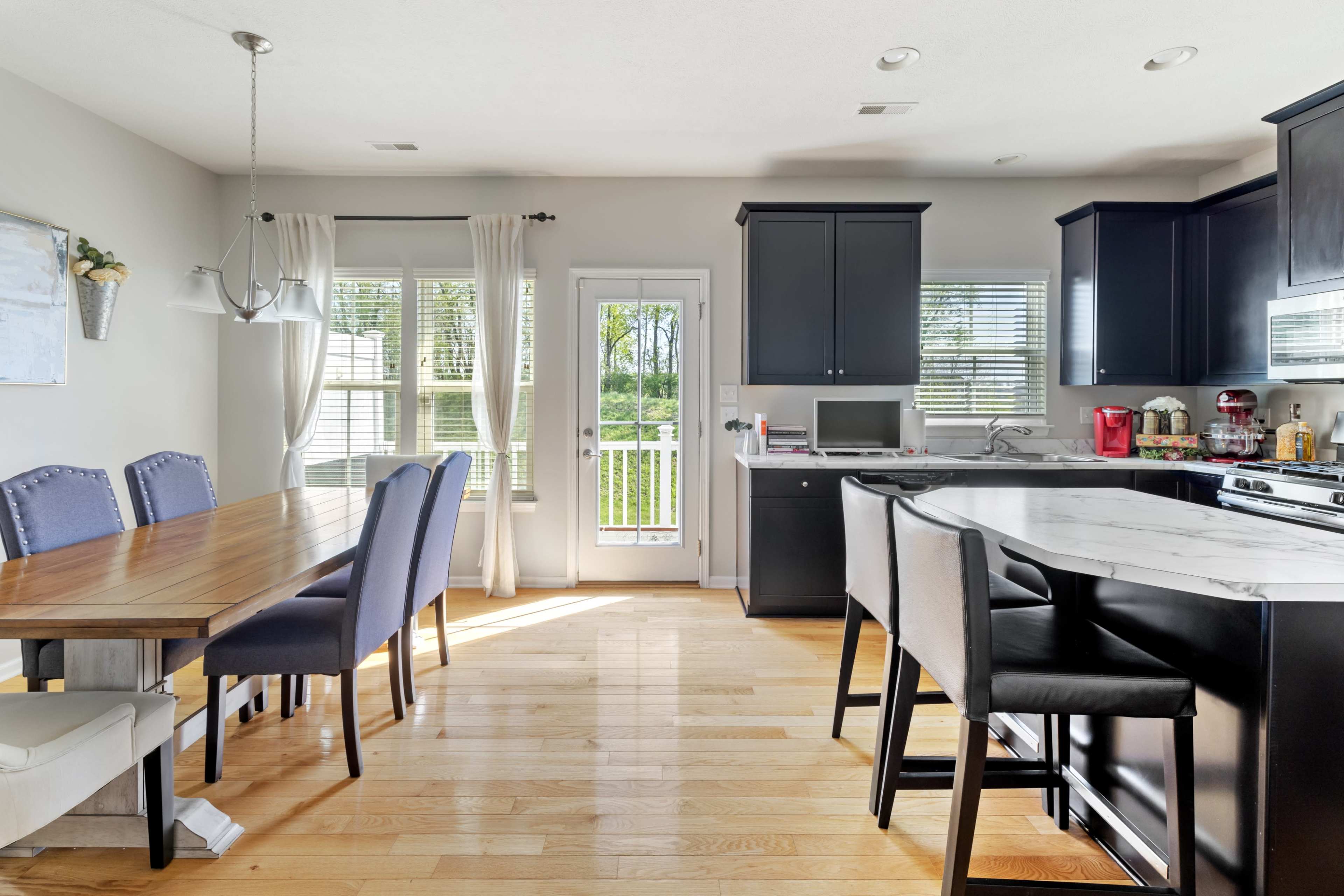 A modern kitchen and dining area features a wooden table with chairs, a kitchen island with bar stools, and large windows overlooking a view.
