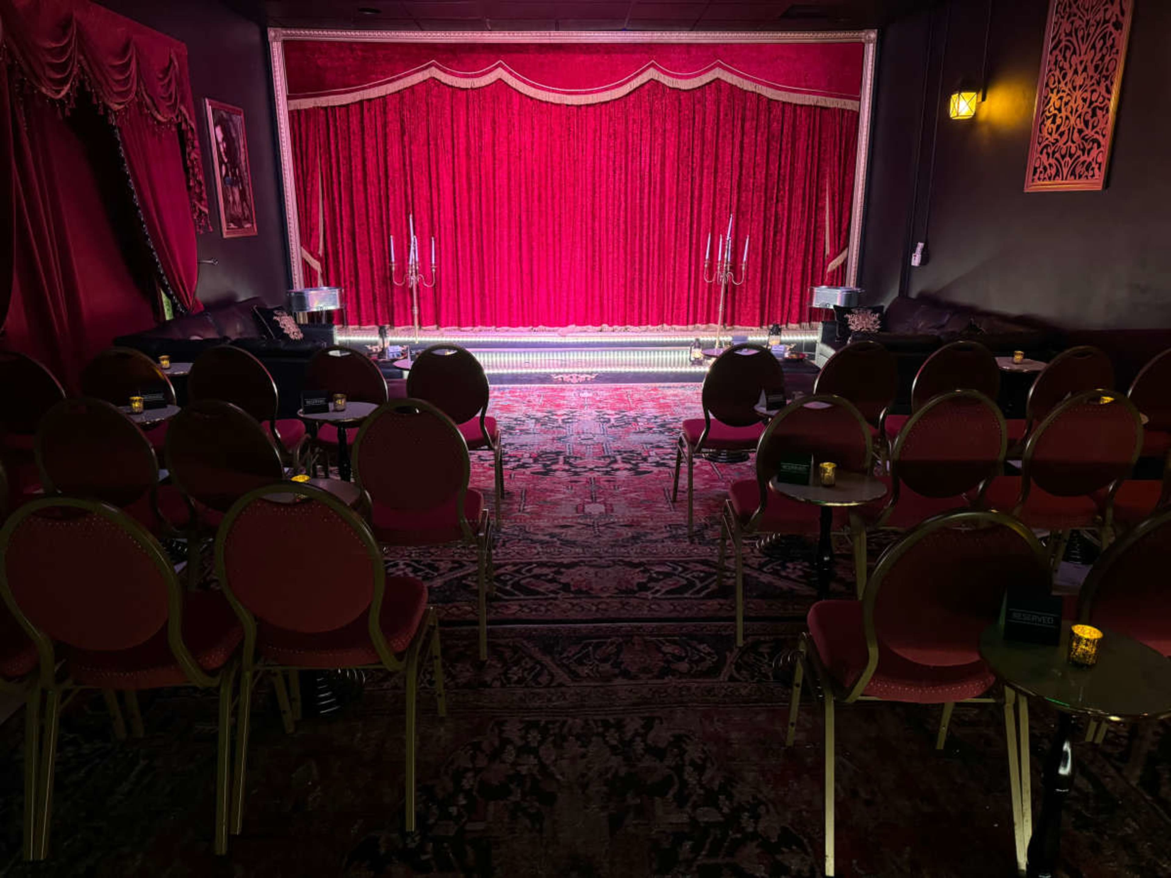 A performing arts venue features a stage draped in red velvet curtains, with empty round tables and chairs arranged in front of it.