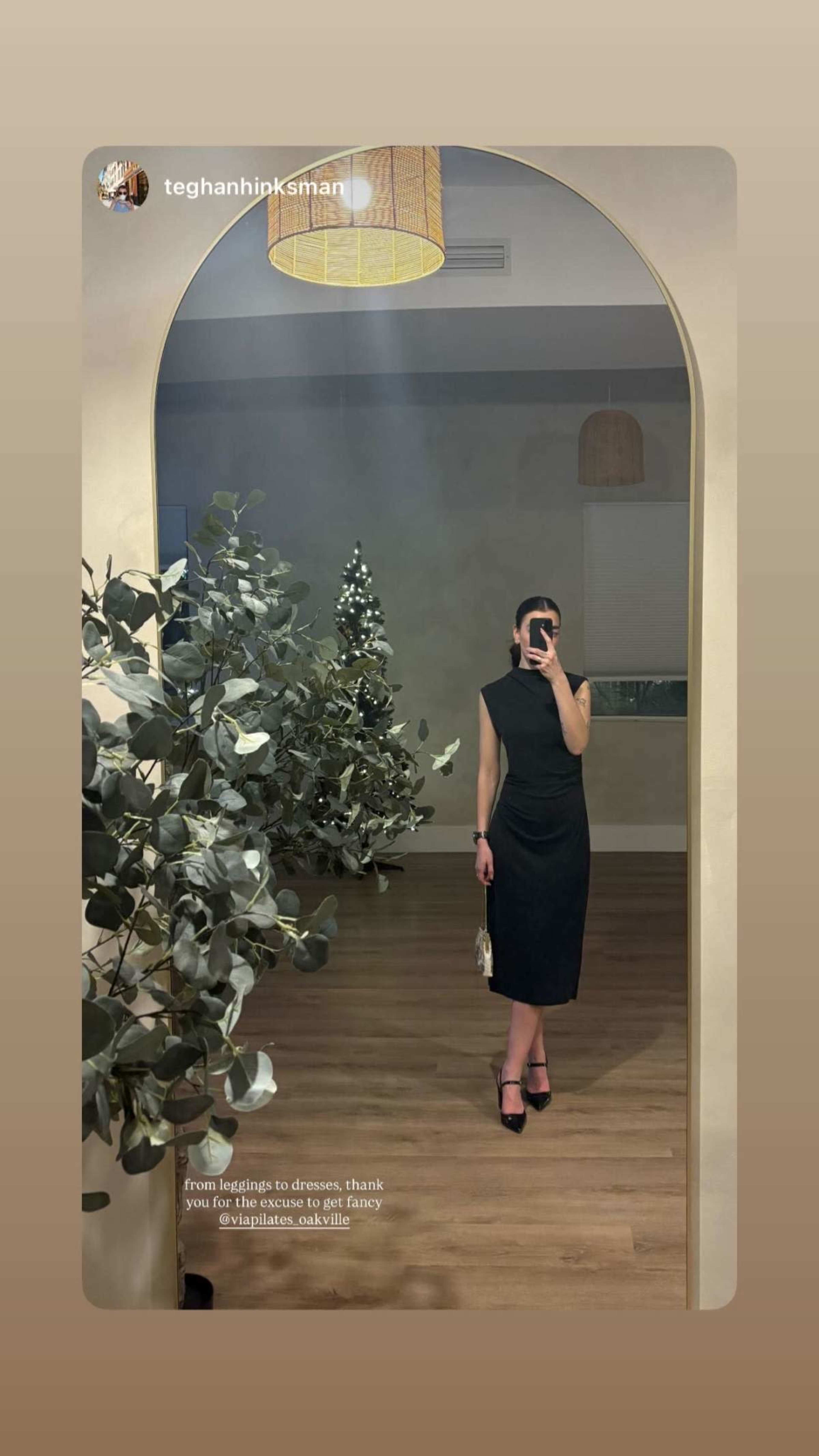 A person stands in front of a large mirror, wearing a fitted black dress and holding a small bag, while decorated Christmas trees are visible in the background.