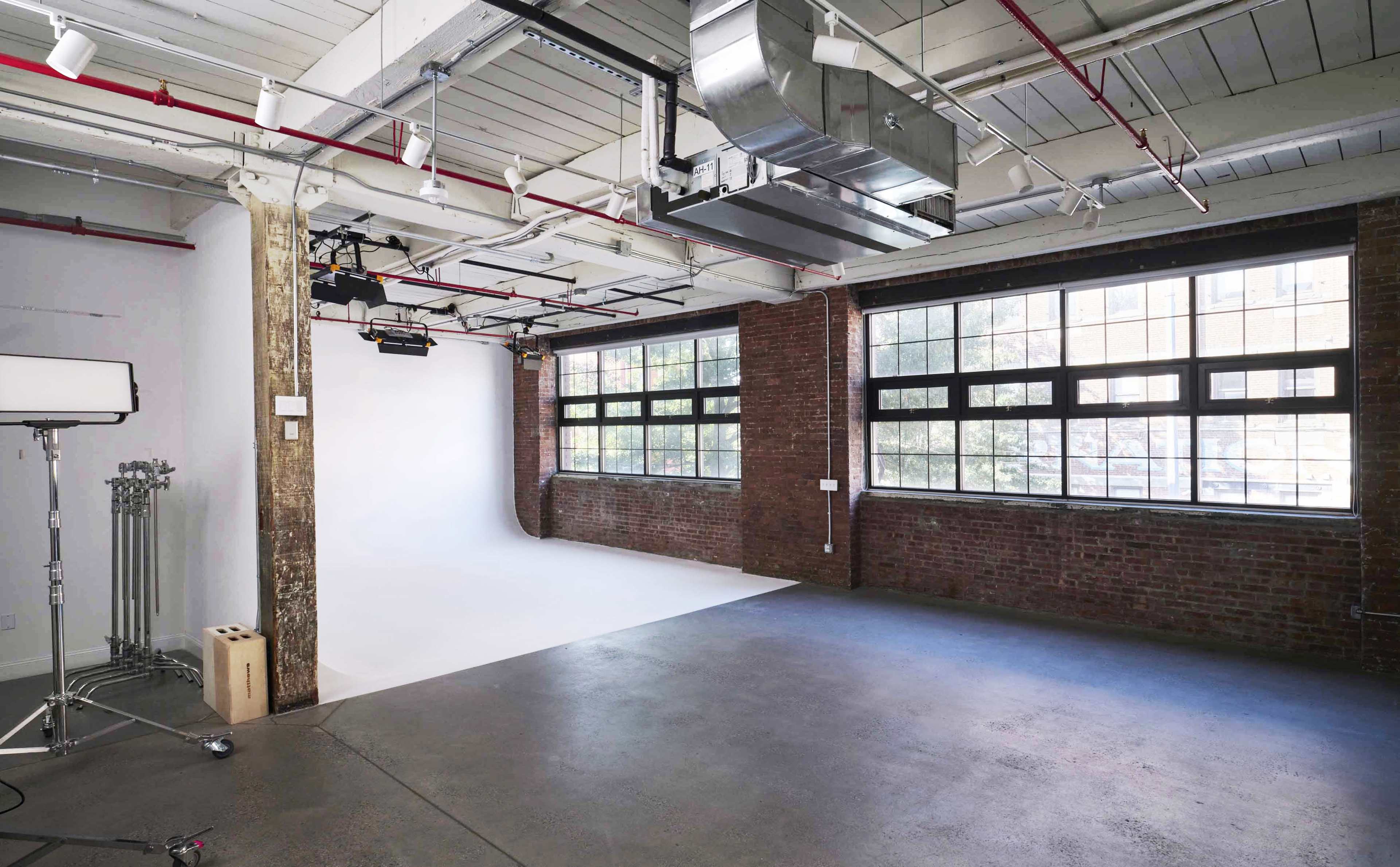 A spacious photography studio with exposed brick walls, large windows, and a white backdrop setup.