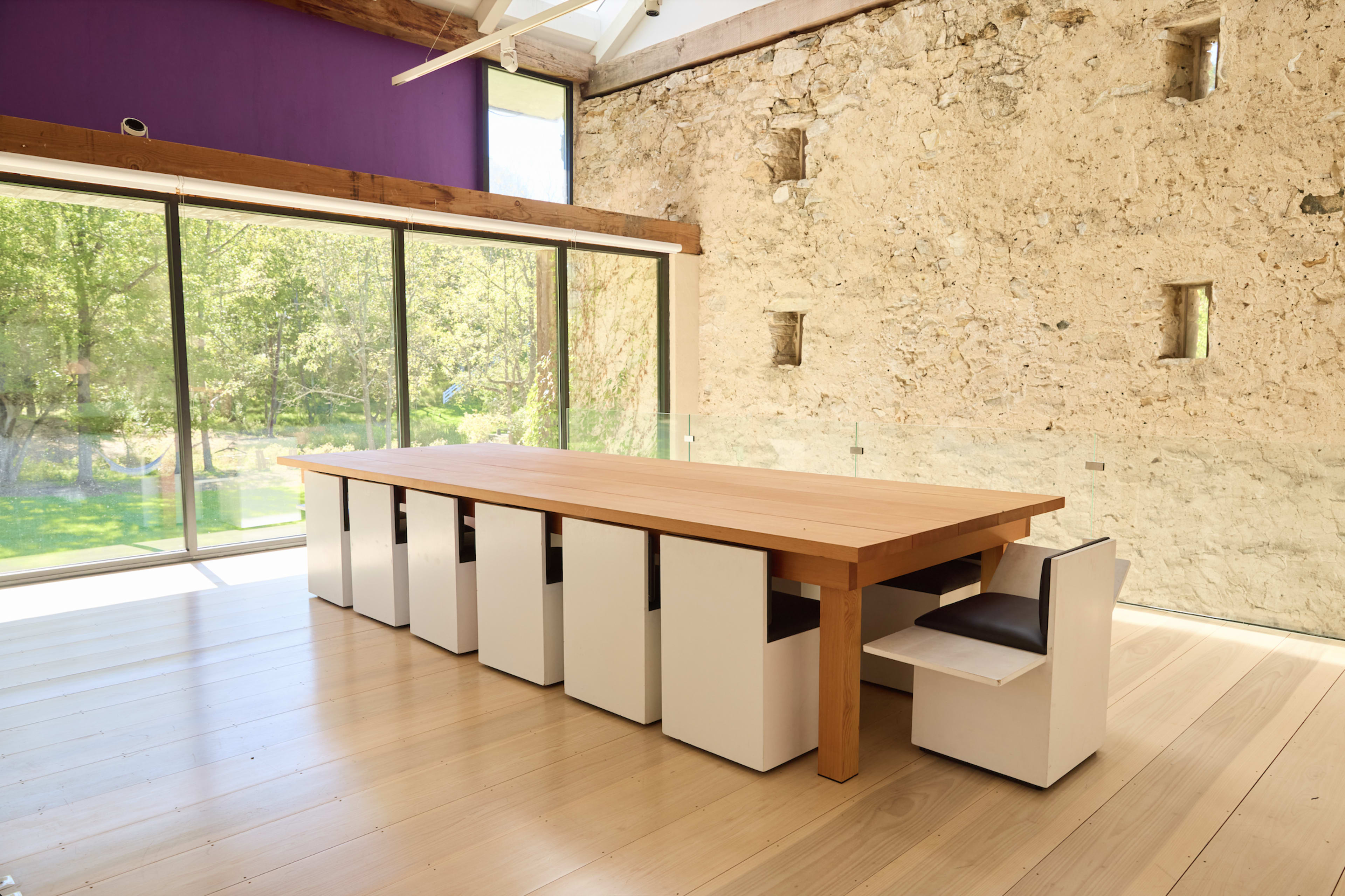 The image shows a spacious room with a large wooden table surrounded by minimalist white chairs, and a glass wall revealing a view of greenery outside.