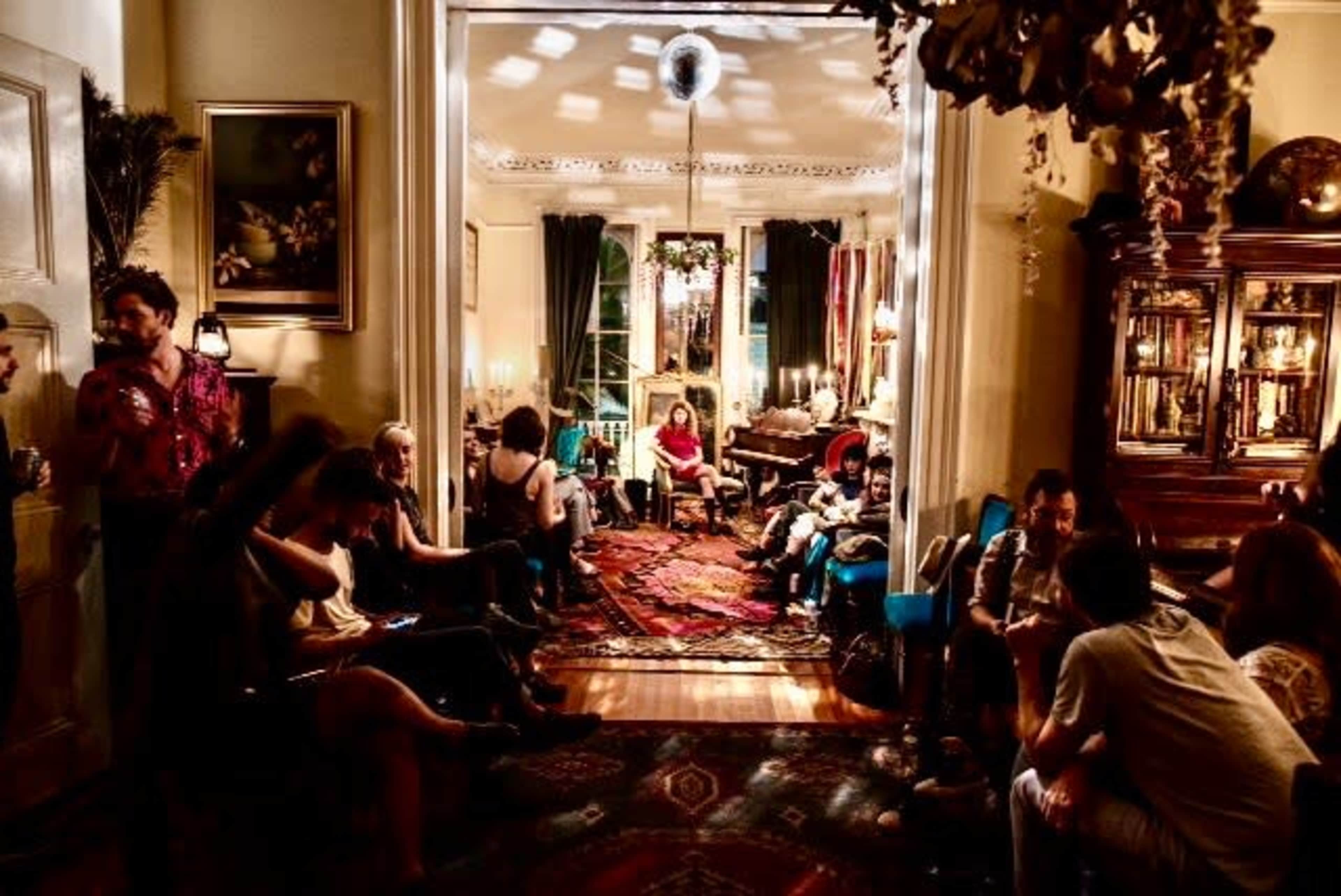 A group of people is gathered in a warmly lit living room filled with vintage furniture and decorative items, creating a social atmosphere.