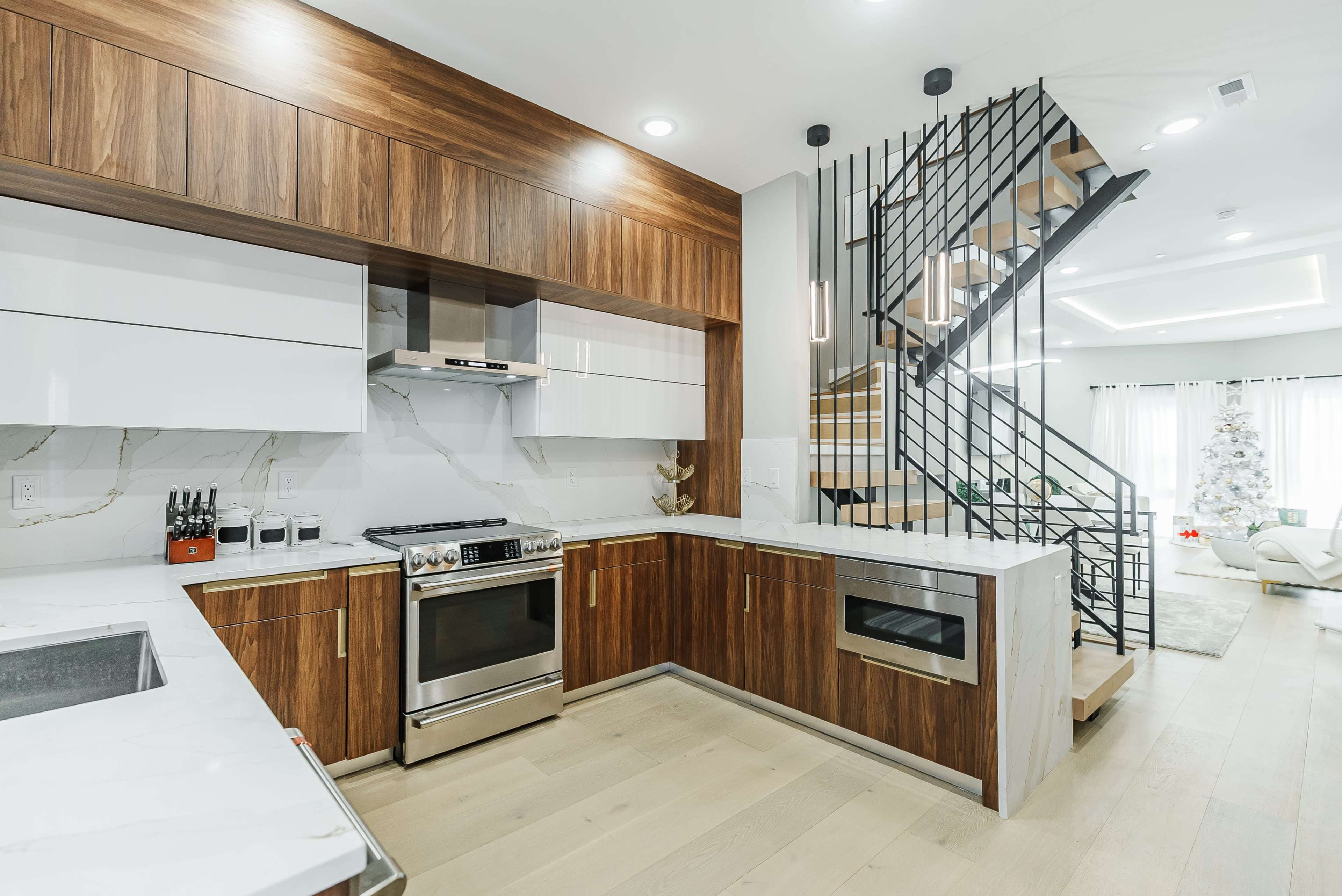 The image shows a modern kitchen with wooden cabinetry, stainless steel appliances, and a staircase with a black metal railing in the background.