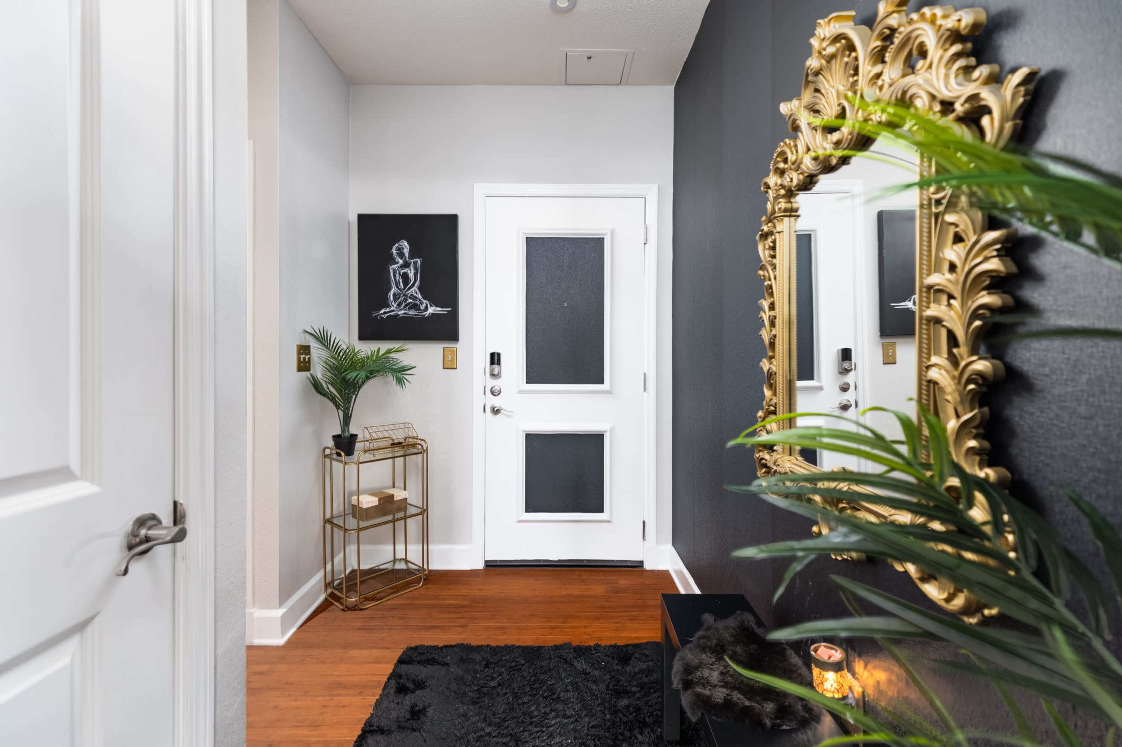 The image shows a stylish entryway featuring a gold-framed mirror, a small decorative table, a potted plant, and a dark area rug against a wall with a contrasting paint color.