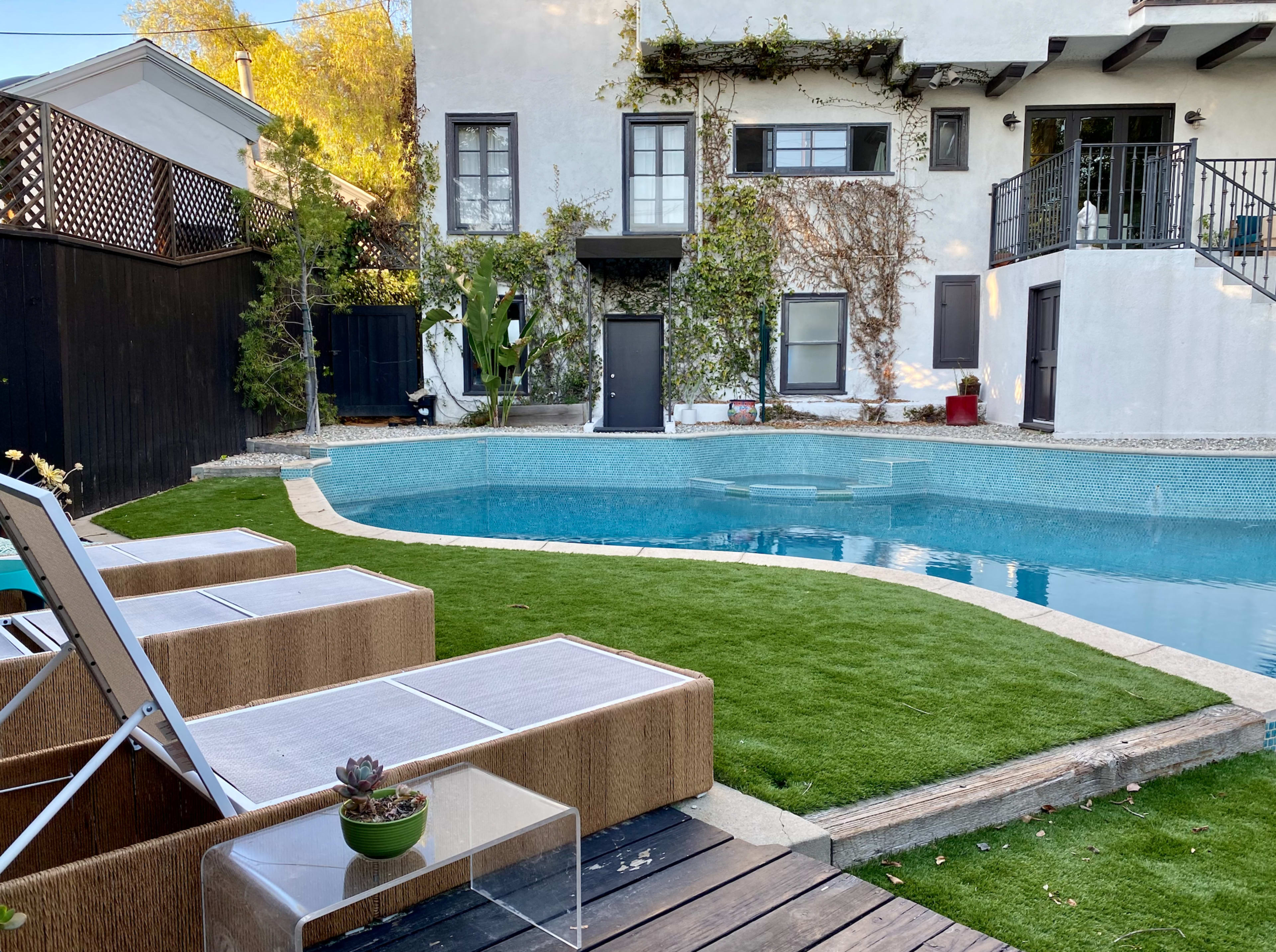 A backyard features a swimming pool surrounded by green grass, with lounge chairs positioned nearby and a white two-story house in the background.