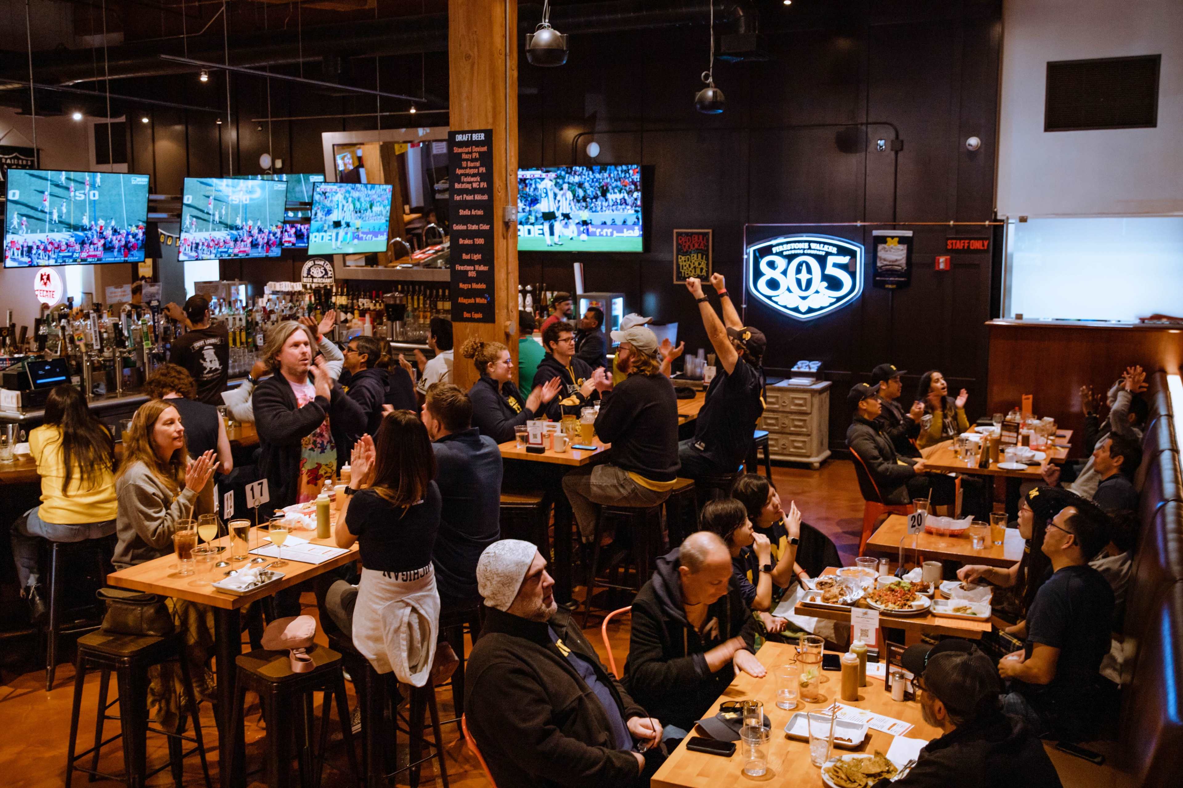 A bustling sports bar is filled with patrons watching games on multiple screens while enjoying food and drinks at wooden tables.