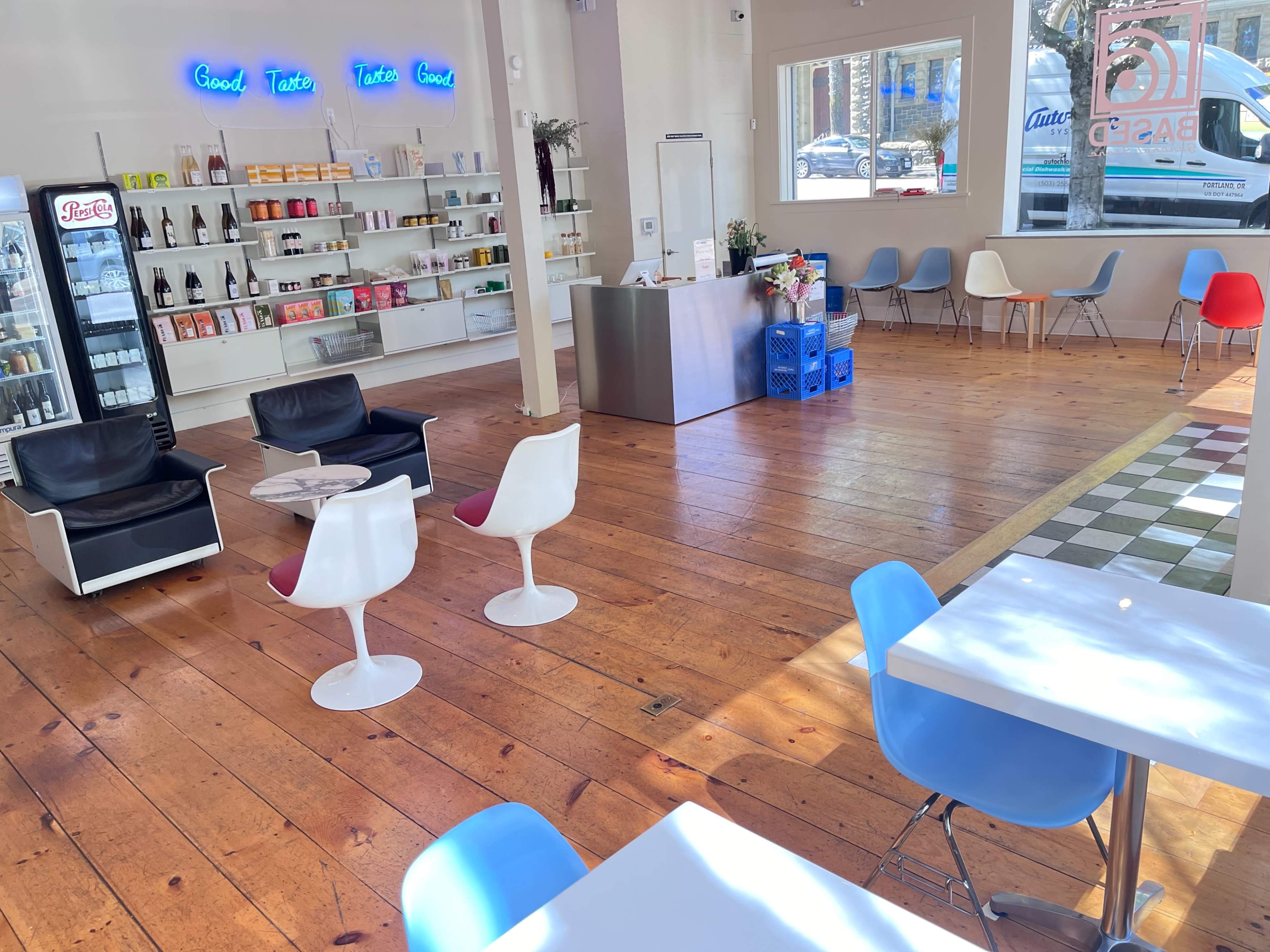 The image shows a bright café interior with wooden flooring, modern seating arrangements, a display shelf of products, and a service counter.