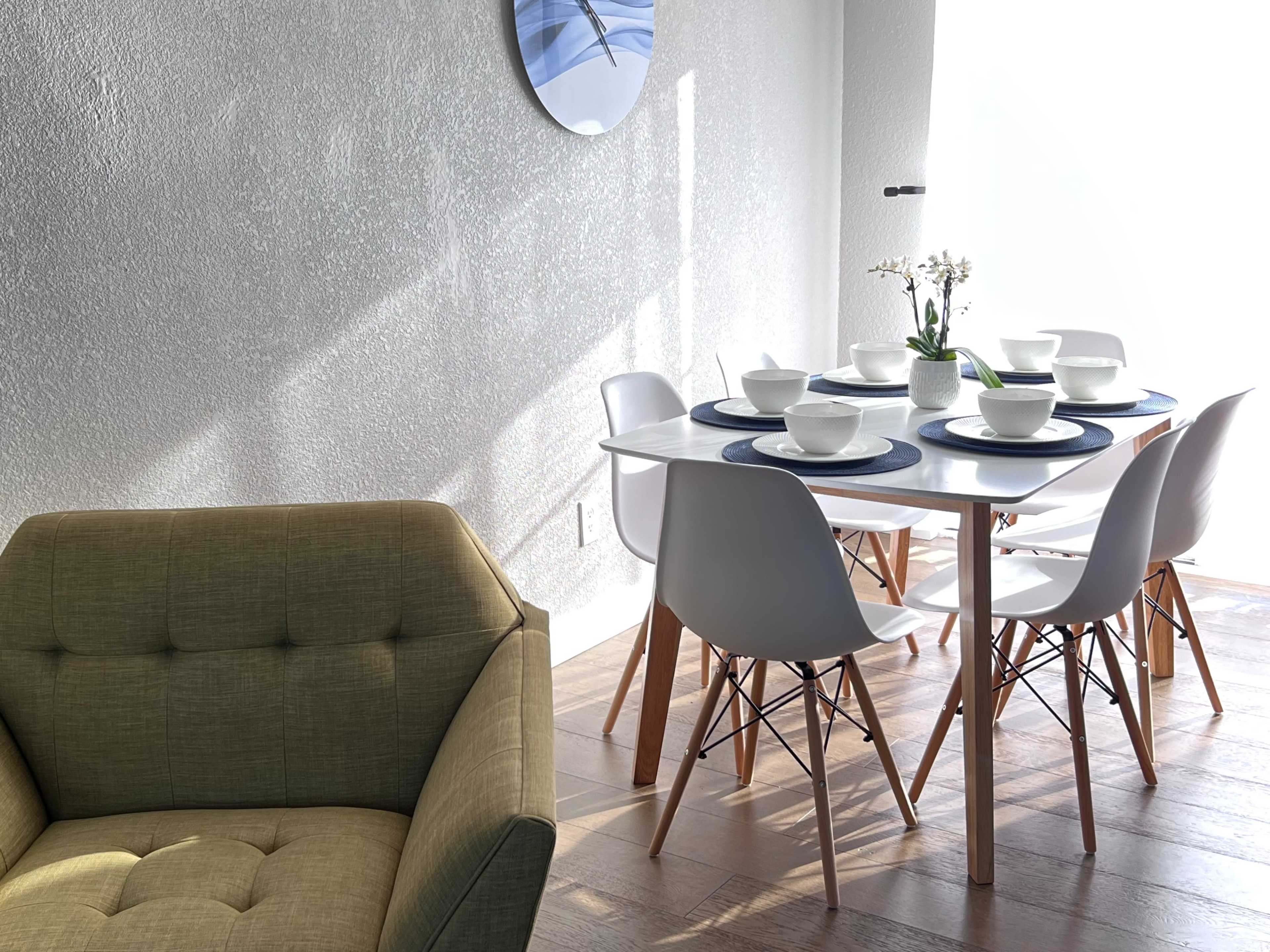 A modern dining area features a white table set for four with bowls on blue placemats, surrounded by white chairs, and complemented by a green upholstered armchair.