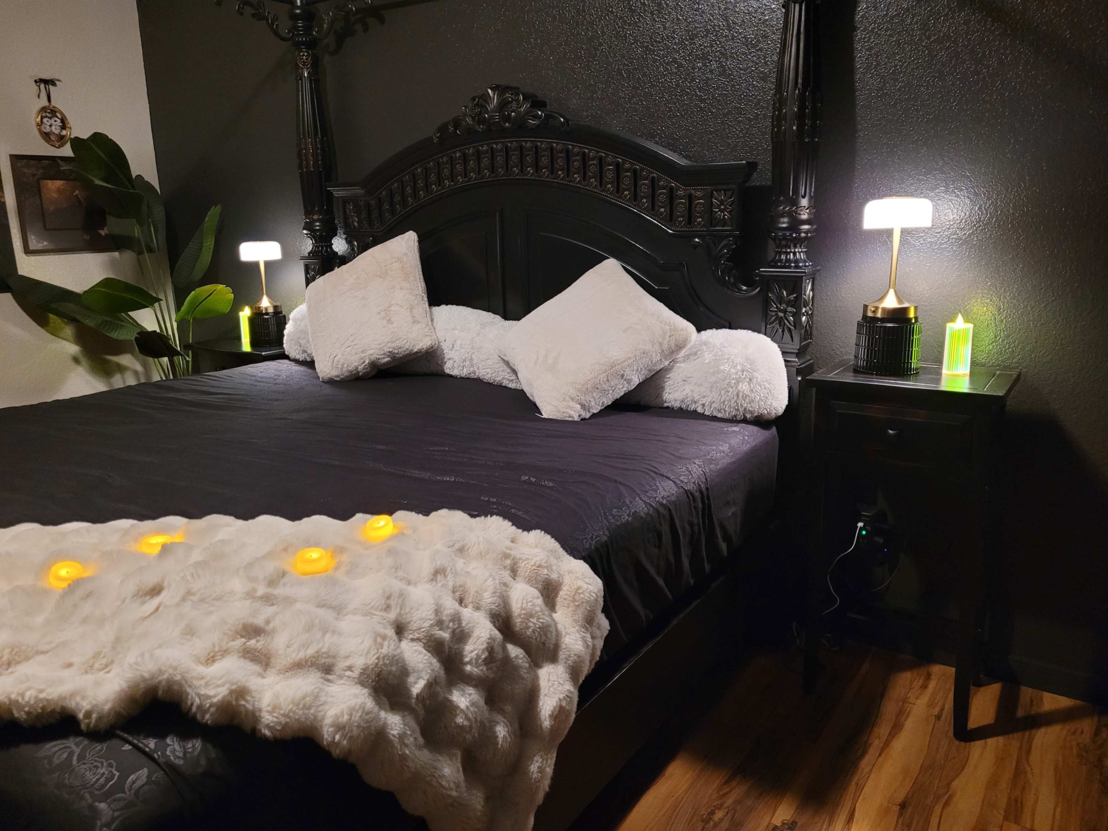 The image shows a dark bedroom featuring a black bed frame with plush pillows, a soft blanket, and bedside lamps casting a warm glow.