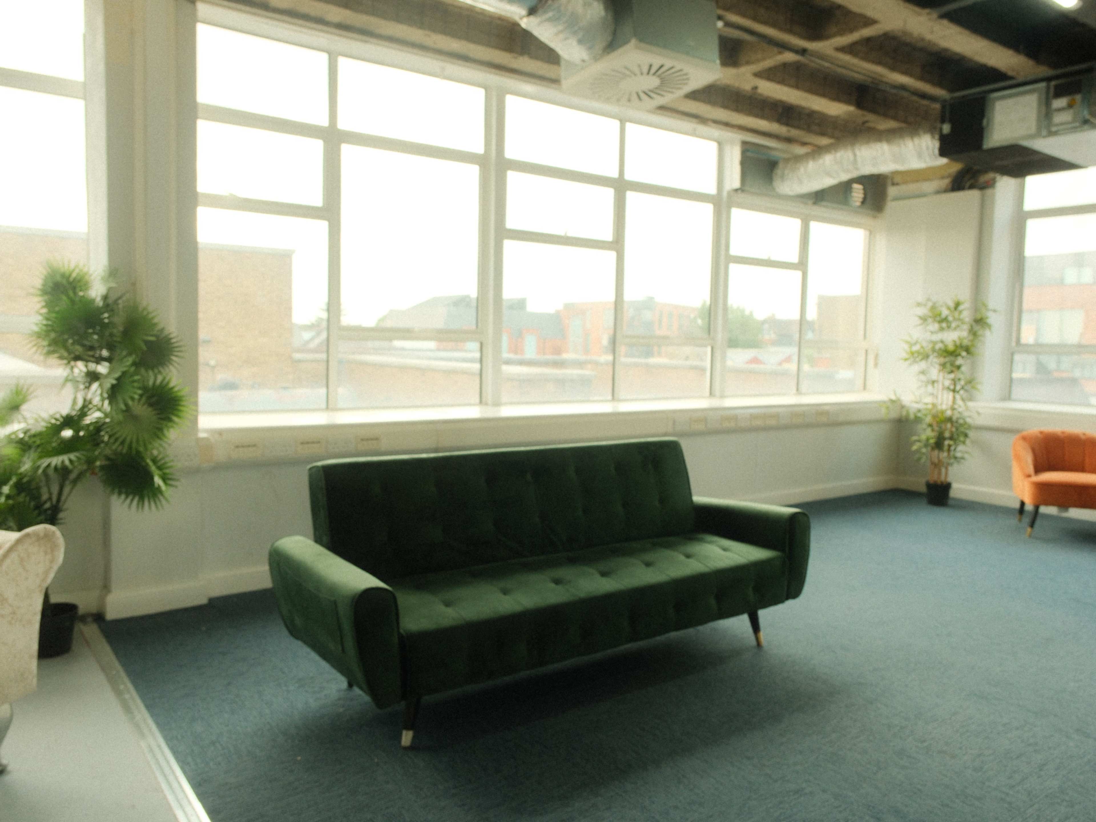 A green sofa is positioned in a bright room with large windows, overlooking a view of rooftops and buildings.
