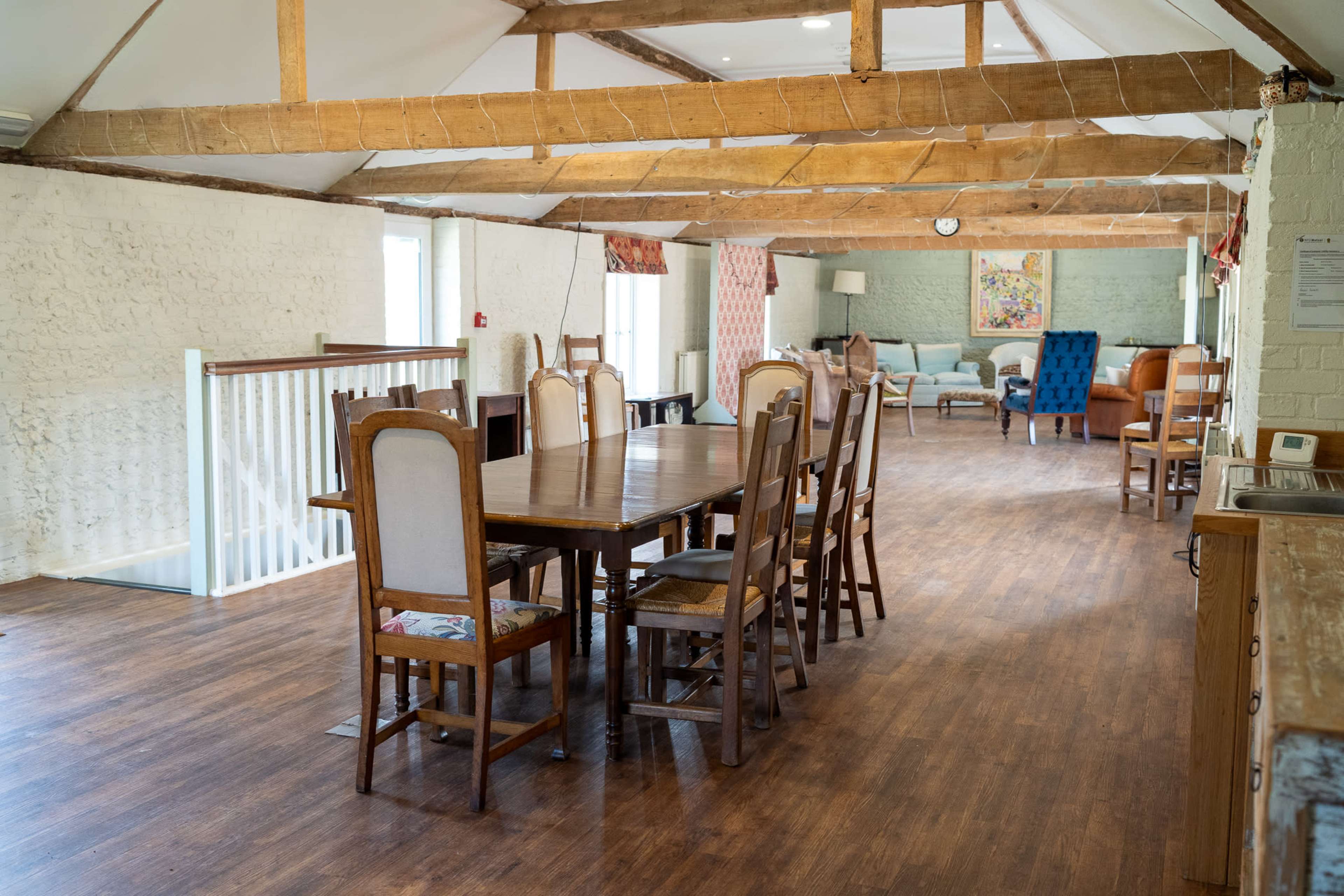 The image shows a spacious interior with wooden beams, featuring a long dining table surrounded by chairs, and various seating areas in a bright, open layout.