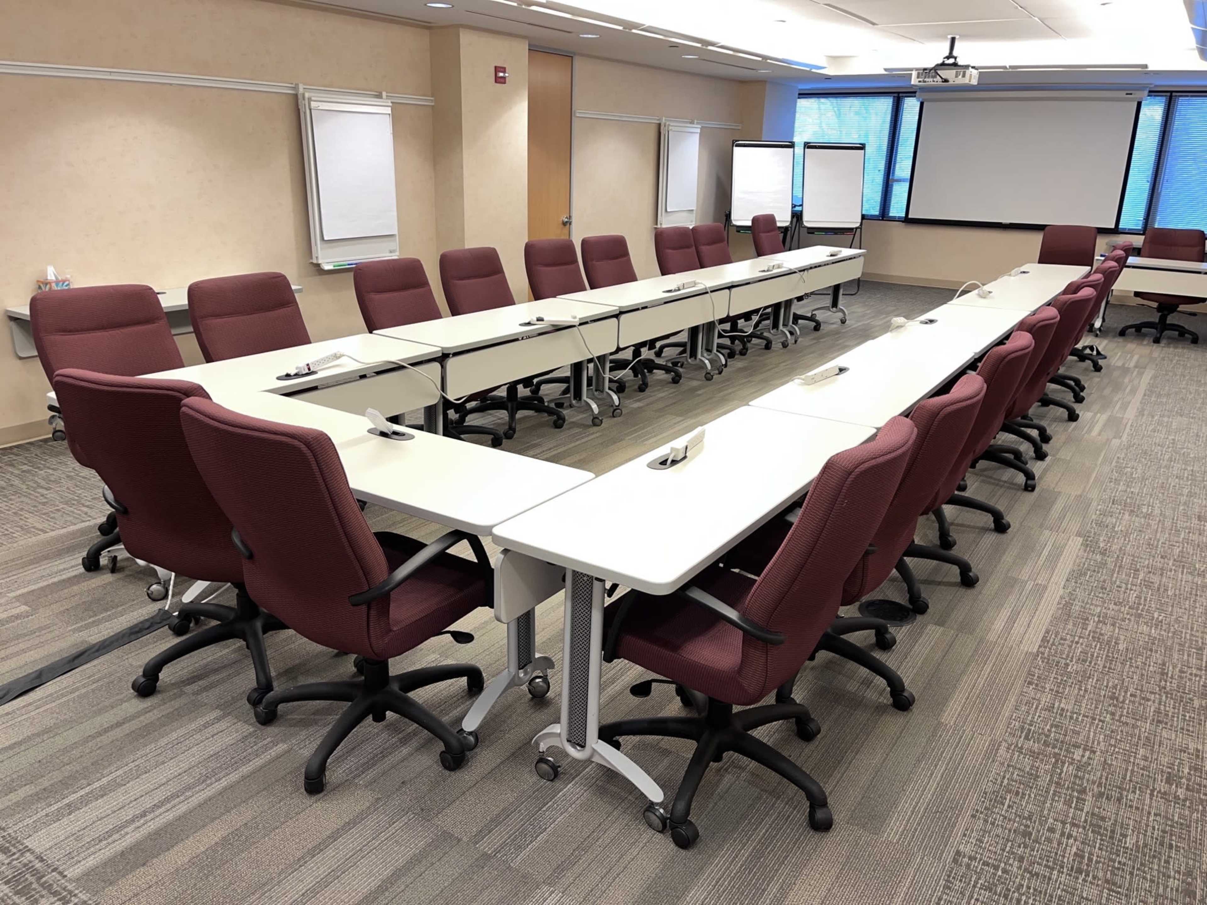 The image shows a conference room with a long, rectangular table surrounded by burgundy chairs and equipped with presentation screens and writing surfaces.