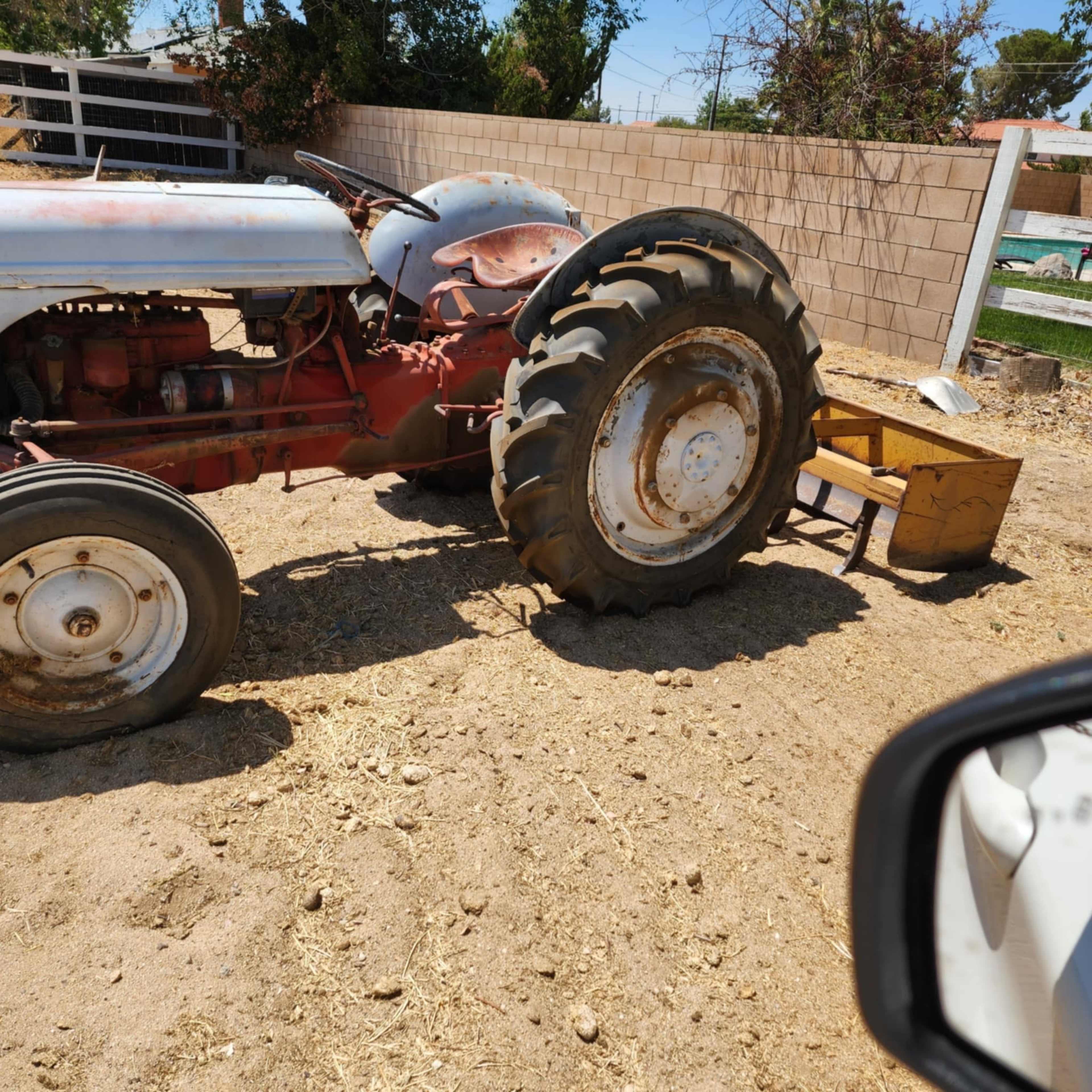 A red vintage tractor with a flat rear tire sits in a dusty, rural area next to a yellow implement on the ground.