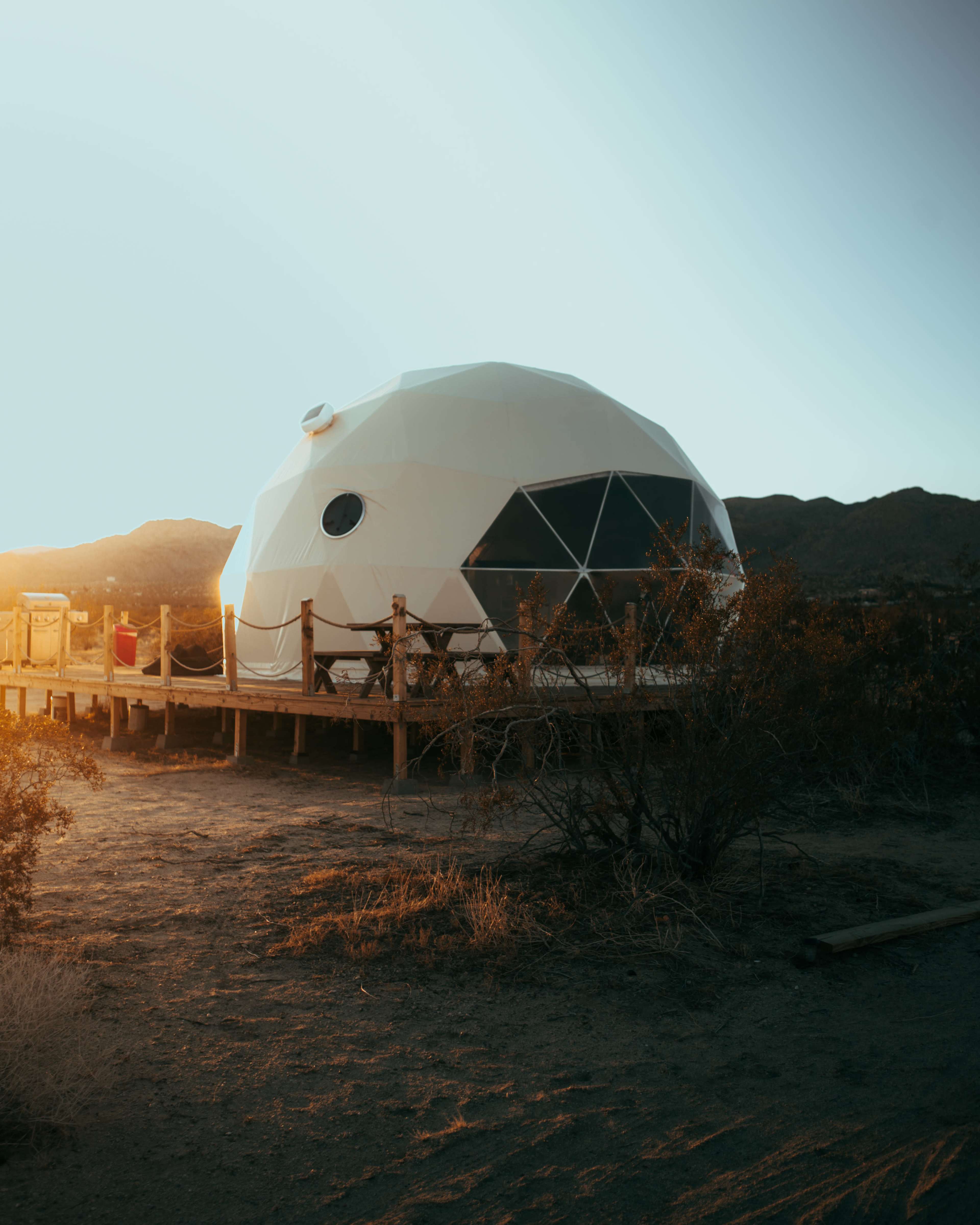 A geodesic dome structure is situated on a wooden deck in a desert landscape with mountains in the background during sunset.