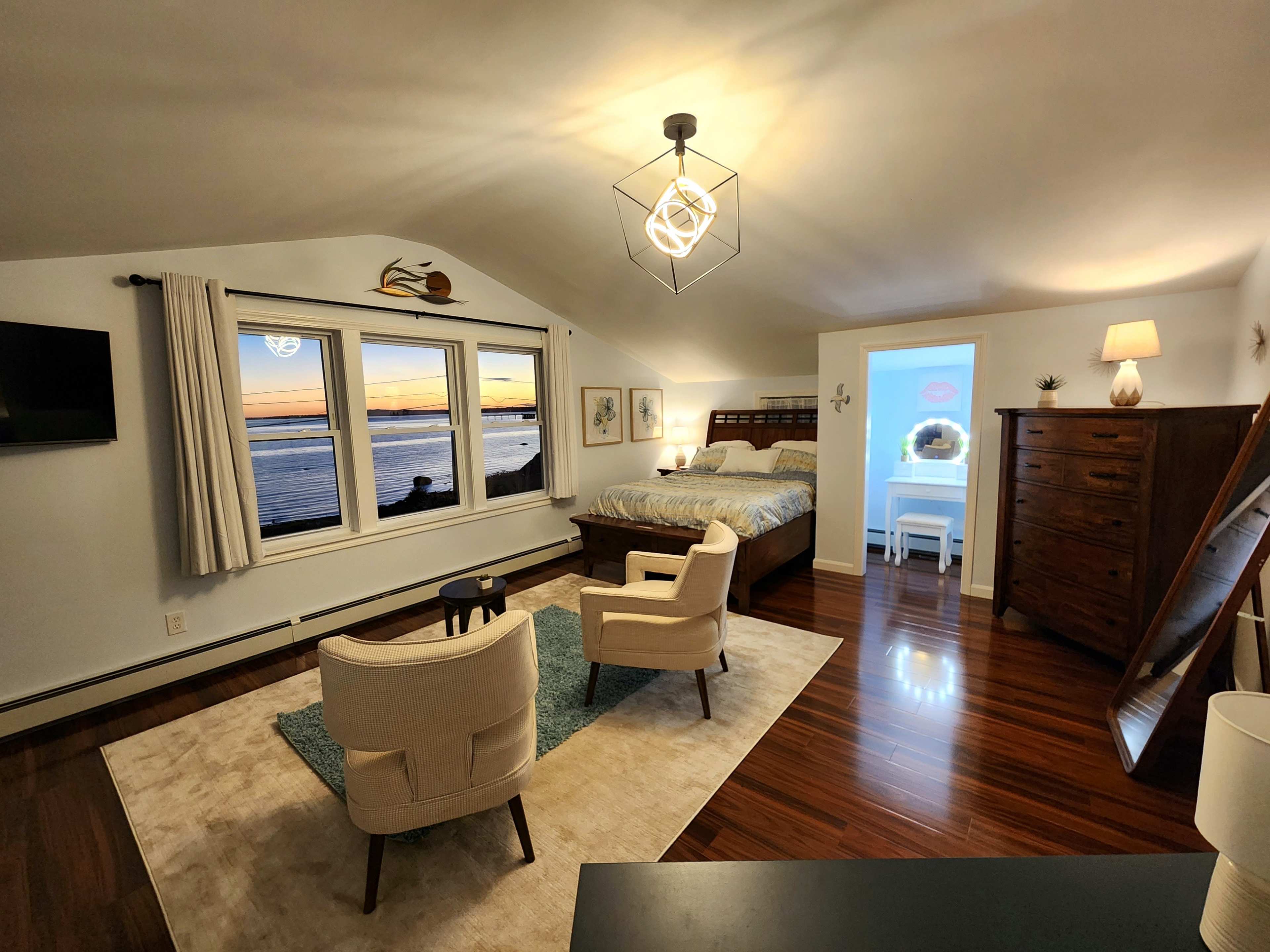A spacious bedroom features a large bed, two chairs on a rug, a dresser, and large windows overlooking a sunset view by the water.