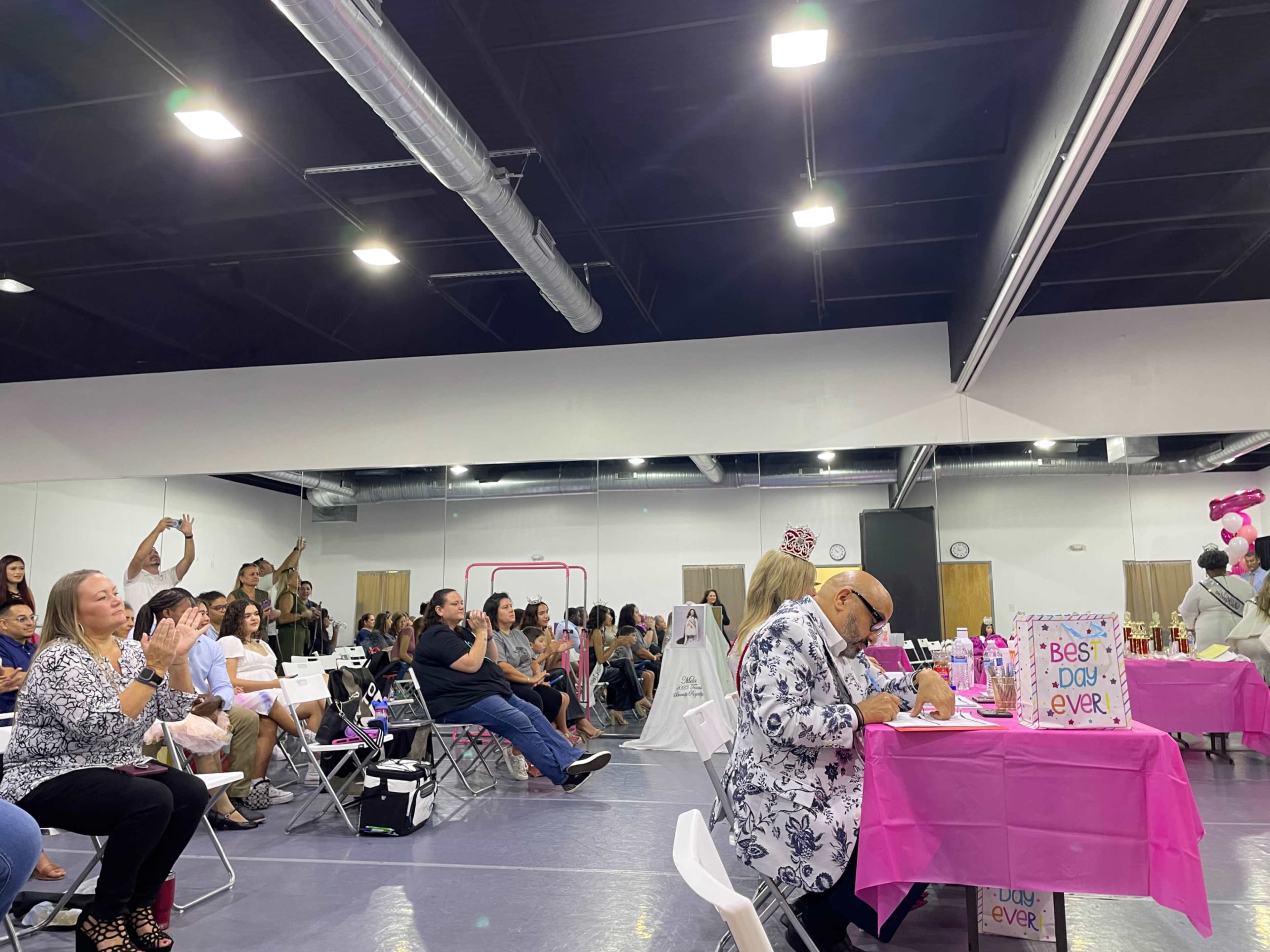 A crowd of people sits in a large, well-lit room, watching a stage event while some attendees take notes at tables adorned with pink decorations.