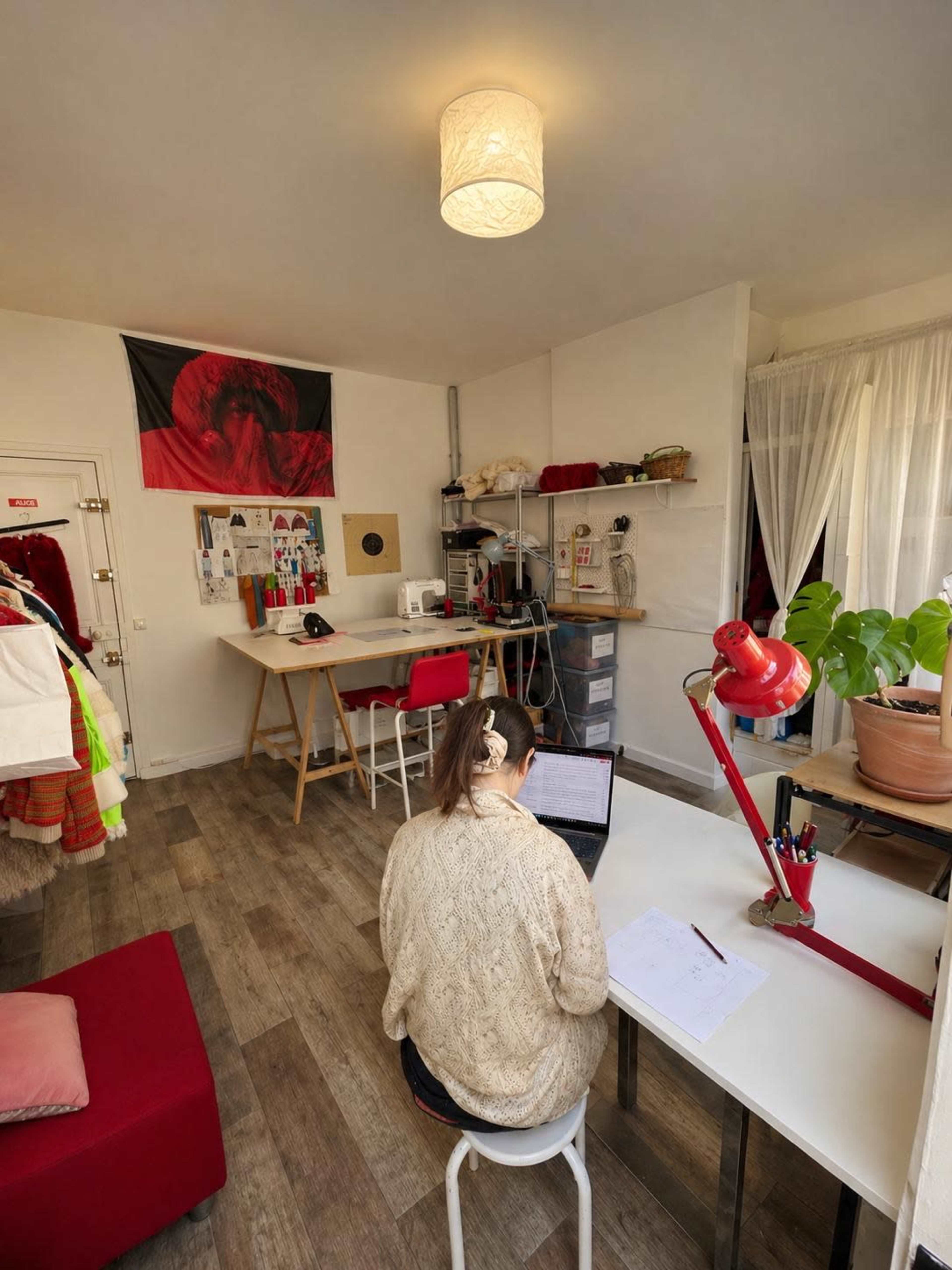 A person sits at a desk working on a laptop in a well-lit room with creative workspace elements and a large wall flag.