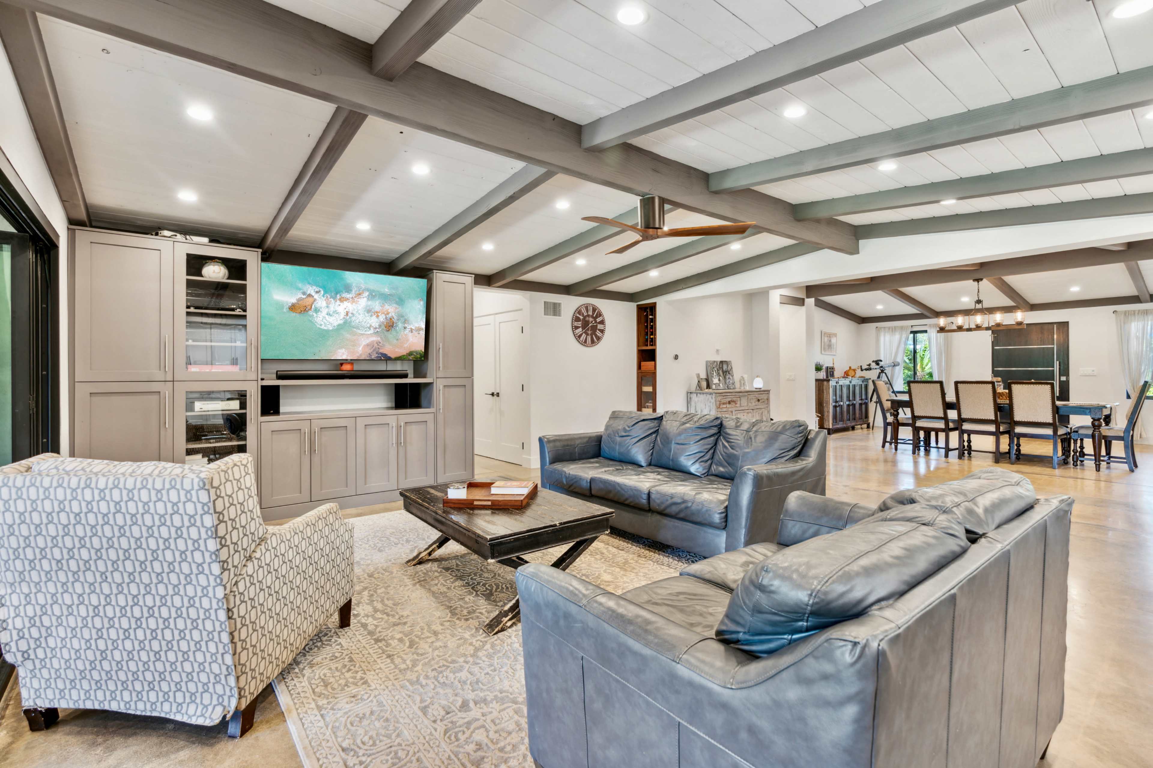 A modern living room with a large flat-screen TV mounted on a wall, two sofas, and a coffee table, all under a beamed ceiling.