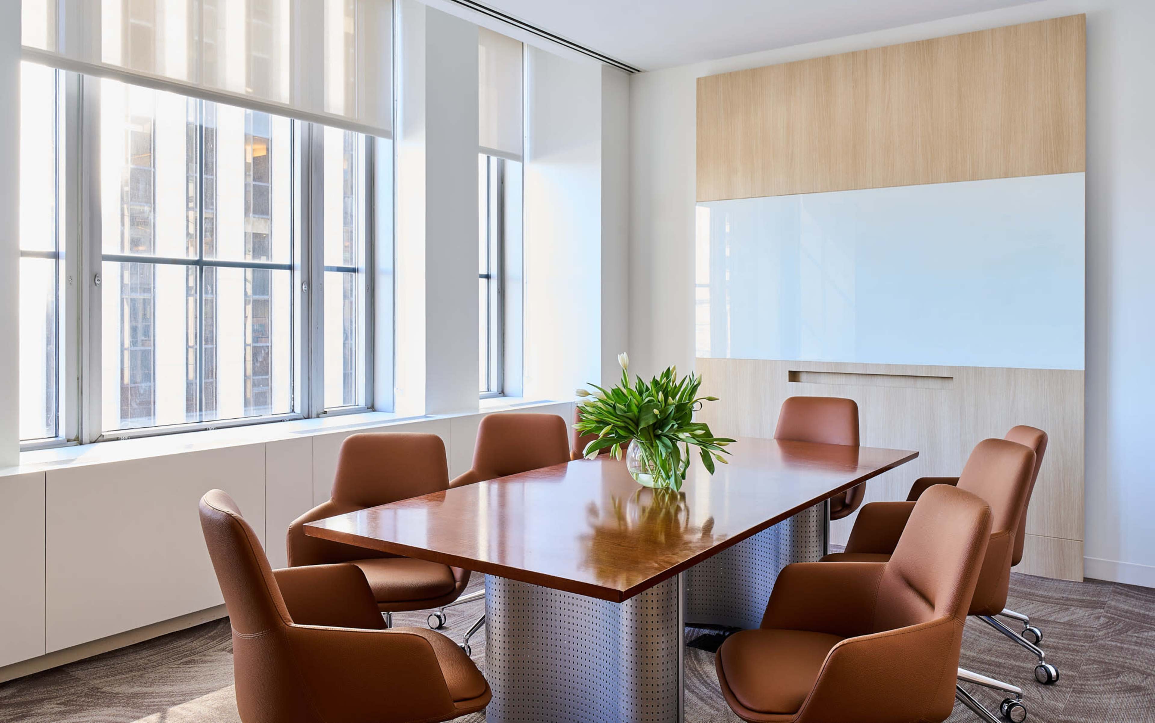 A modern conference room features a large wooden table surrounded by brown leather chairs and a whiteboard on the wall.