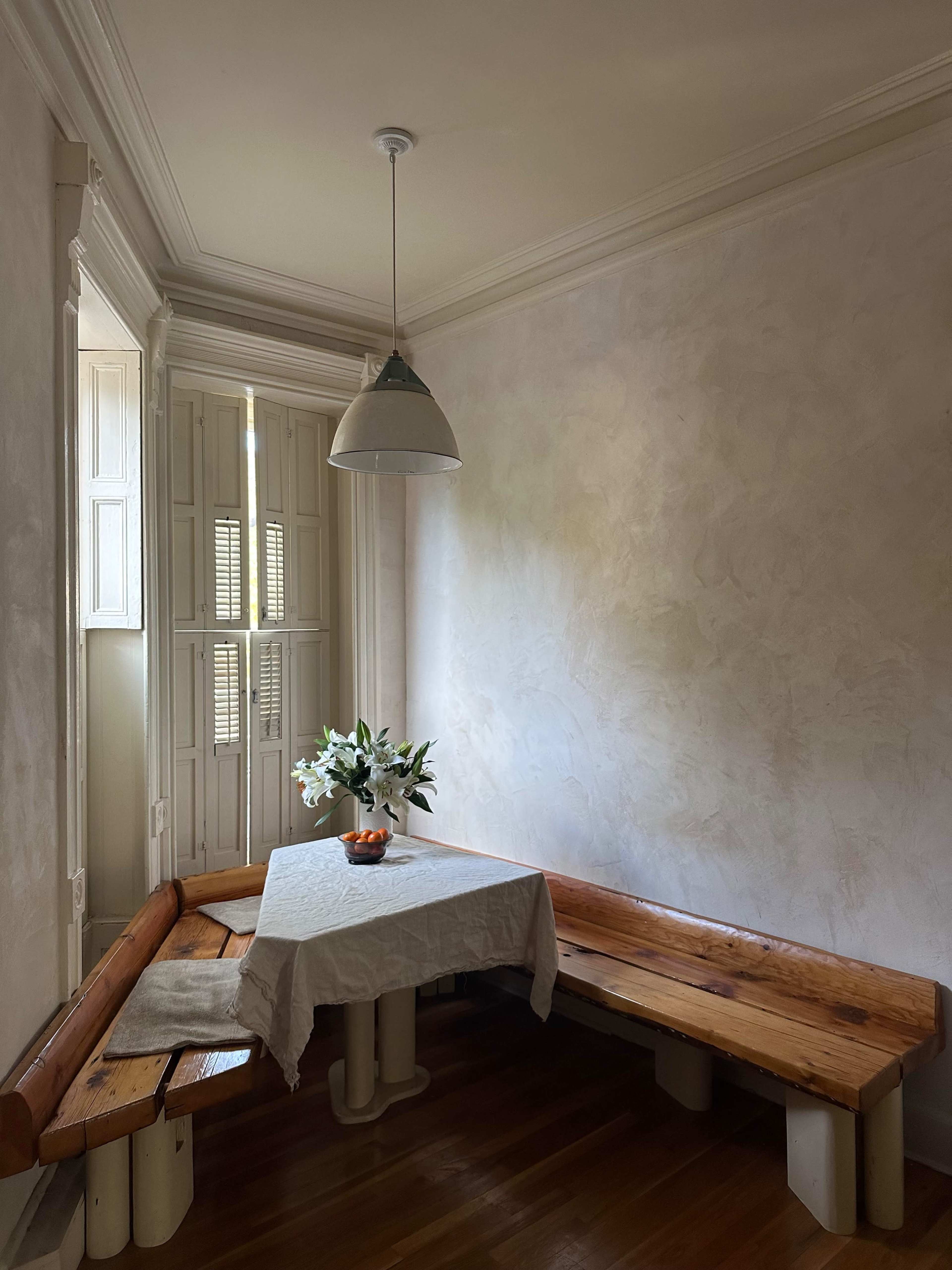 A wooden bench surrounds a small table set with a vase of flowers in a softly lit corner of a room.