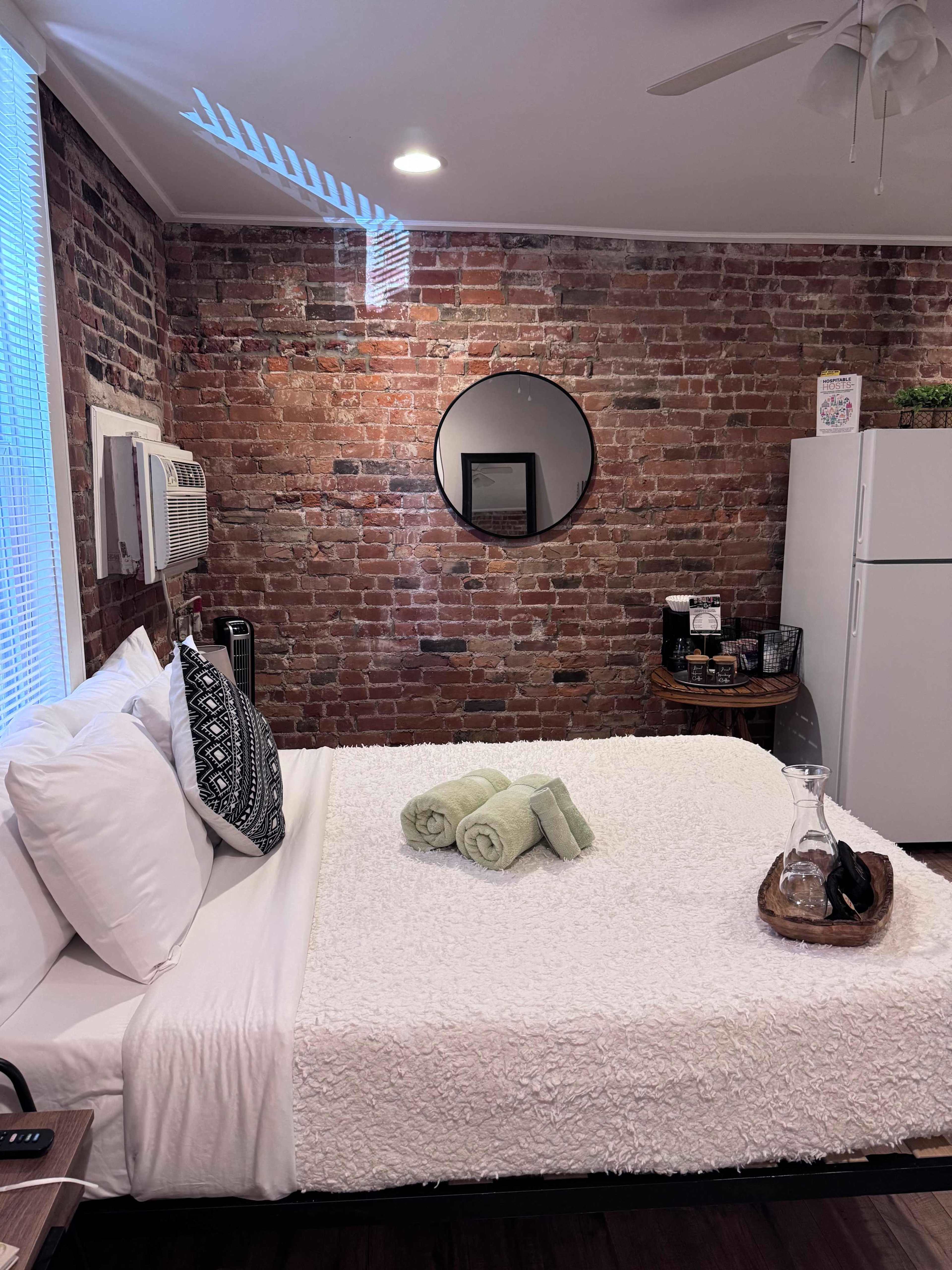 The image shows a cozy bedroom featuring a brick wall, a neatly made bed with white linens, and a round mirror above a small table with towels and decorative items.