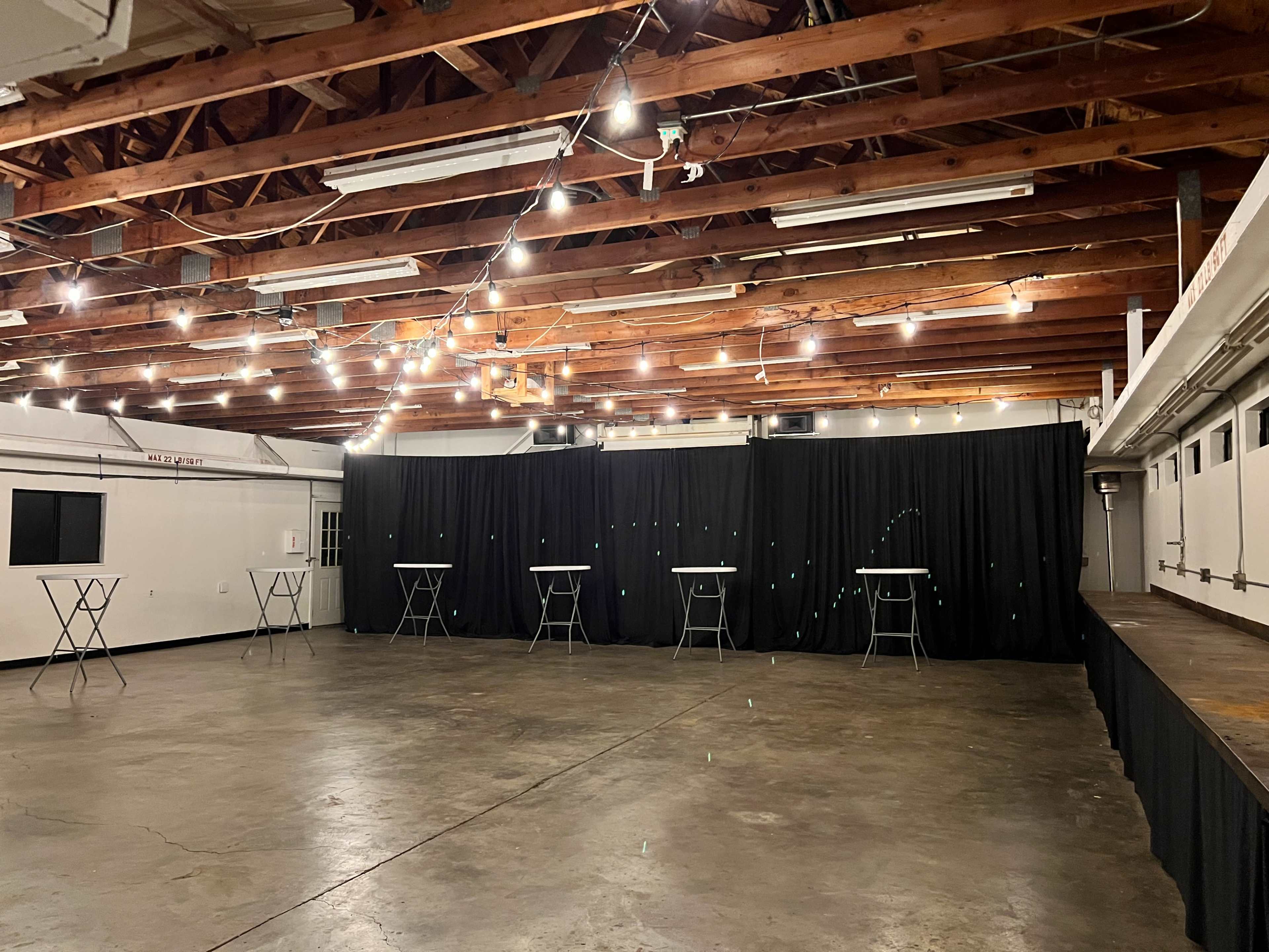 The image shows an empty event space with a concrete floor, folding tables arranged in front of a black curtain, and string lights illuminating the ceiling.