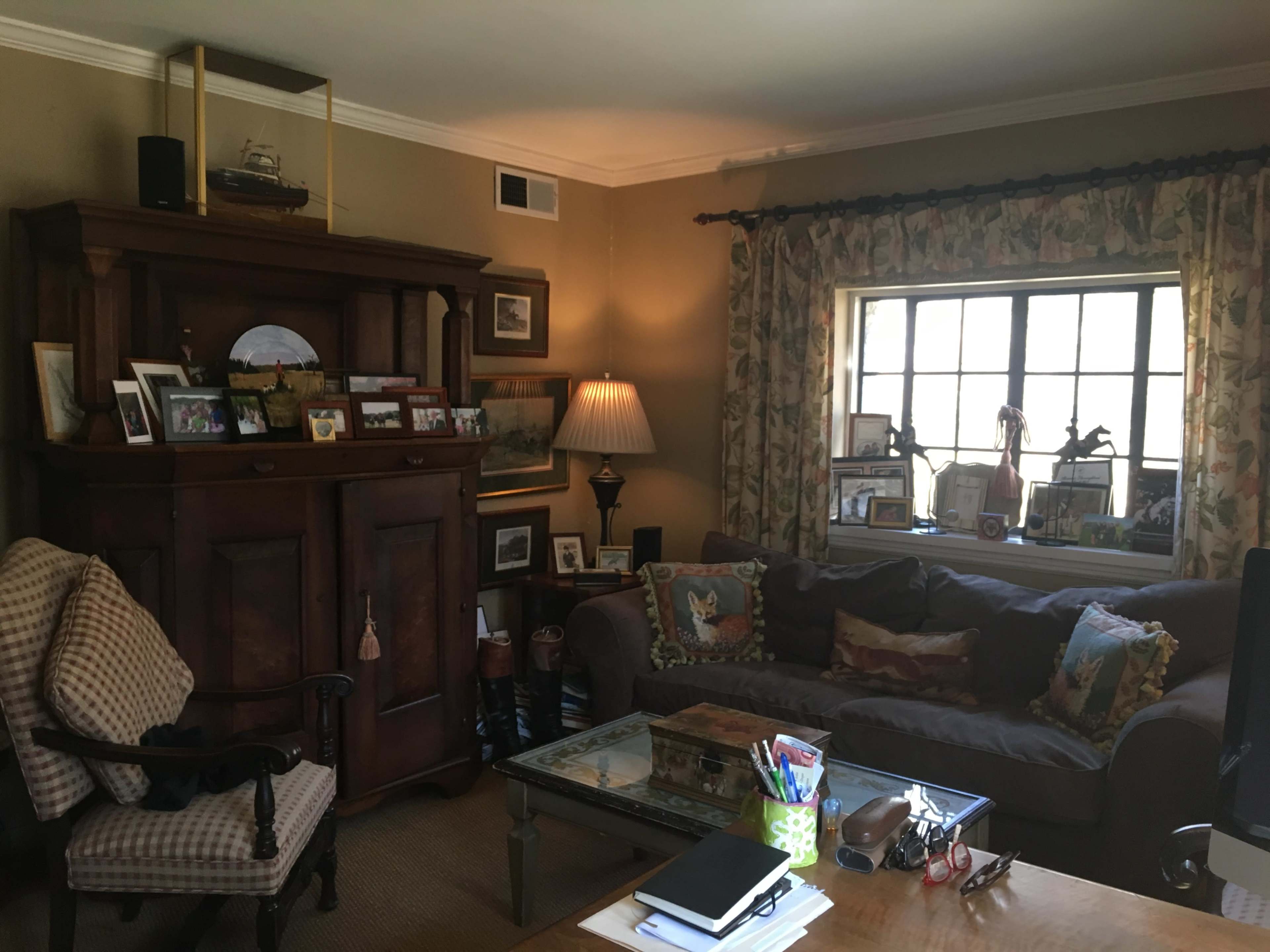 The image shows a cozy living room featuring a dark wooden cabinet, a comfortable sofa with decorative cushions, and a window adorned with patterned curtains, all arranged around a central glass coffee table.