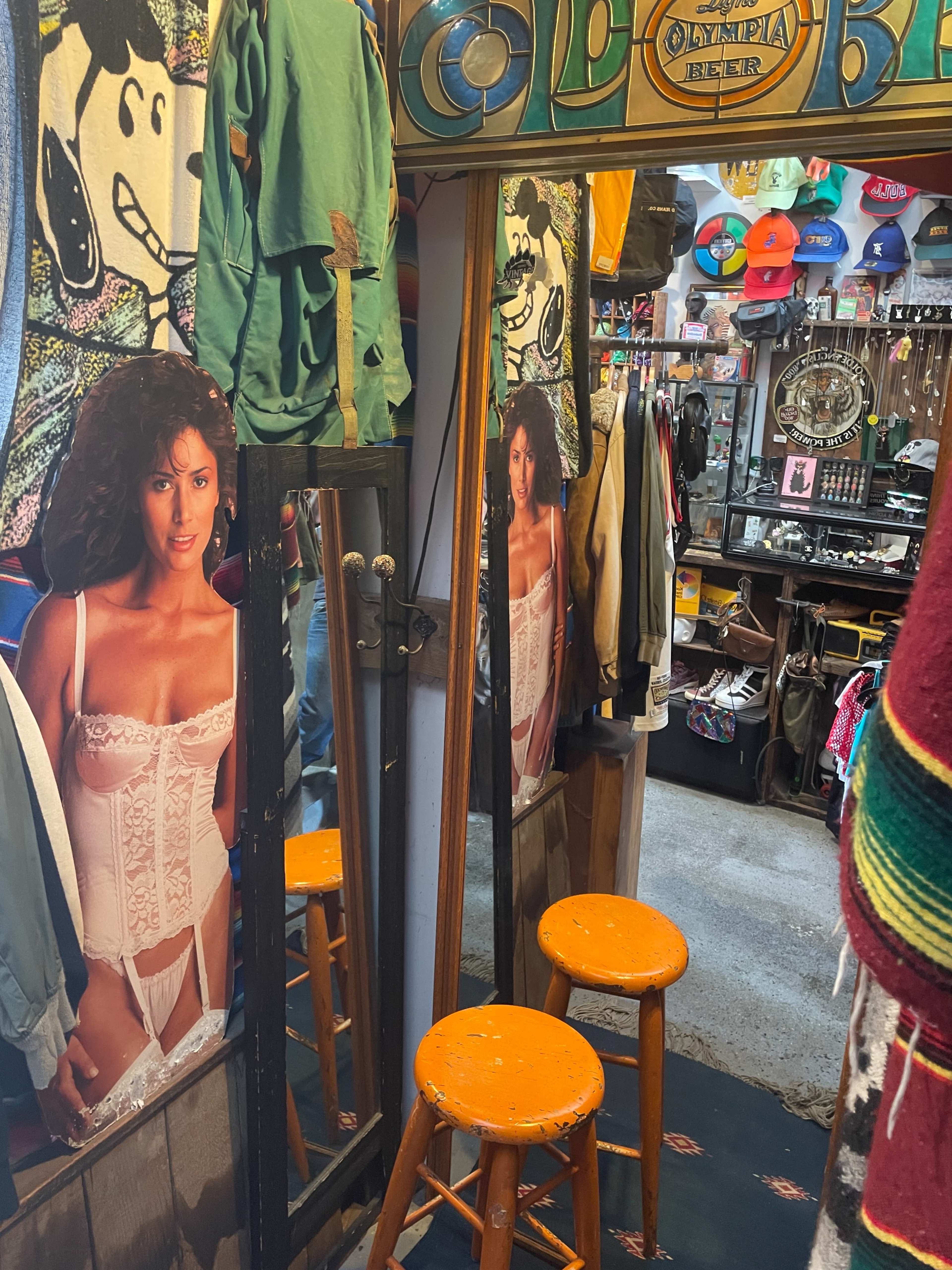 The image shows a colorful vintage clothing store with mirrors reflecting posters of a model, along with wooden stools and various clothing items hanging around.