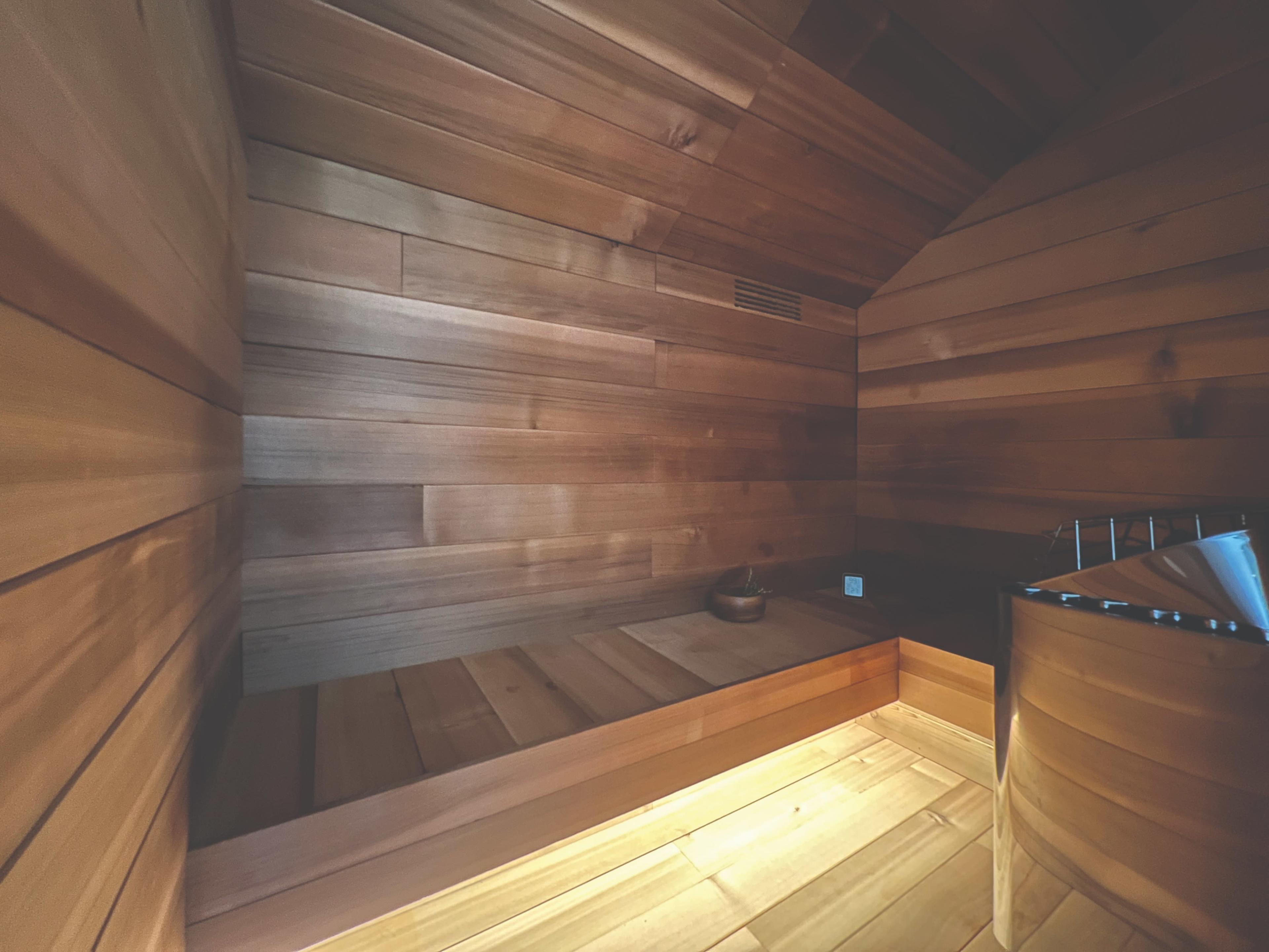 The image shows a wooden sauna with walls and seating made of smooth, light-colored wood and a metal stove in the corner.