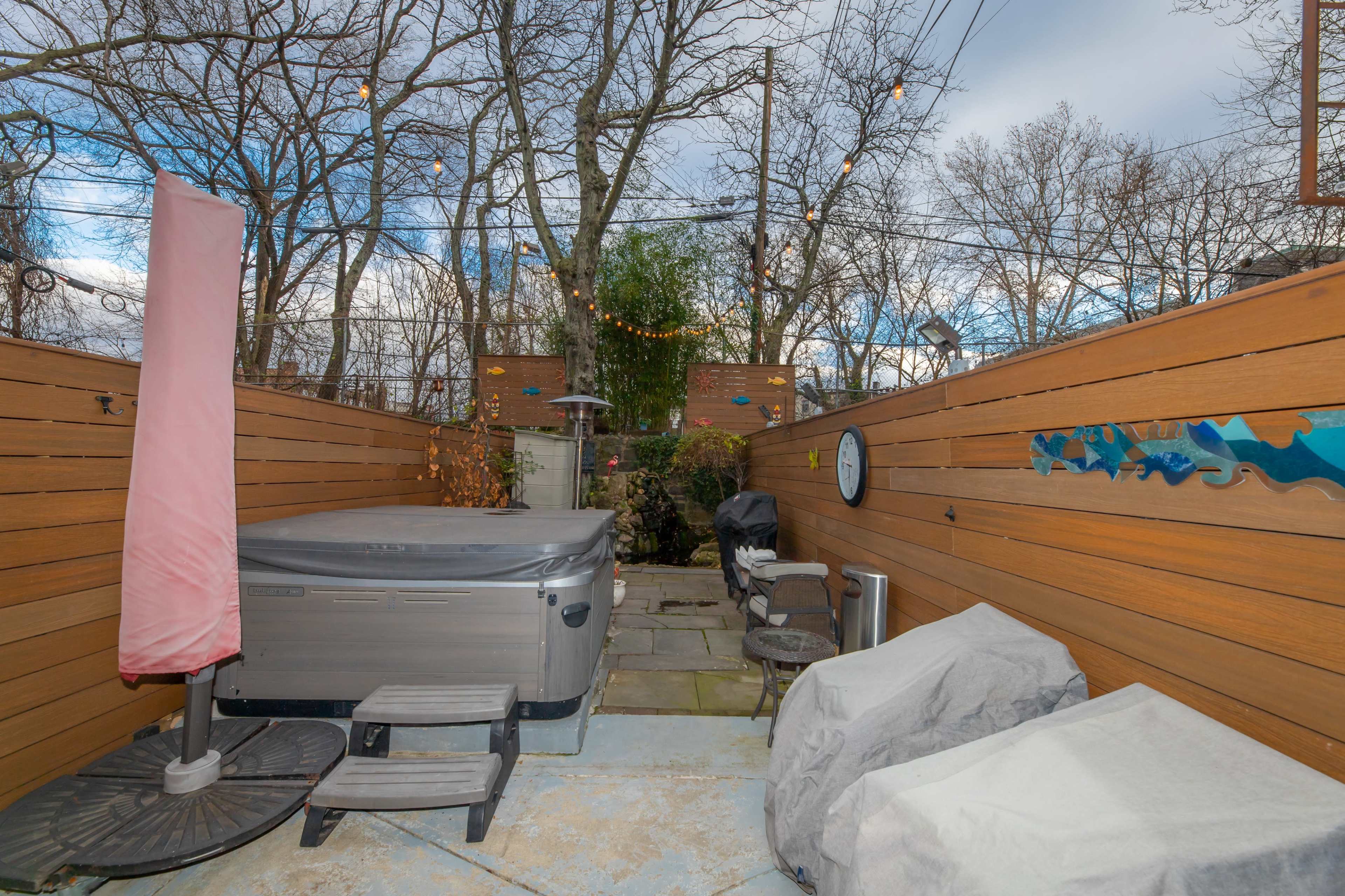 A fenced outdoor space featuring a hot tub, patio furniture, and decorative string lights amidst bare trees.