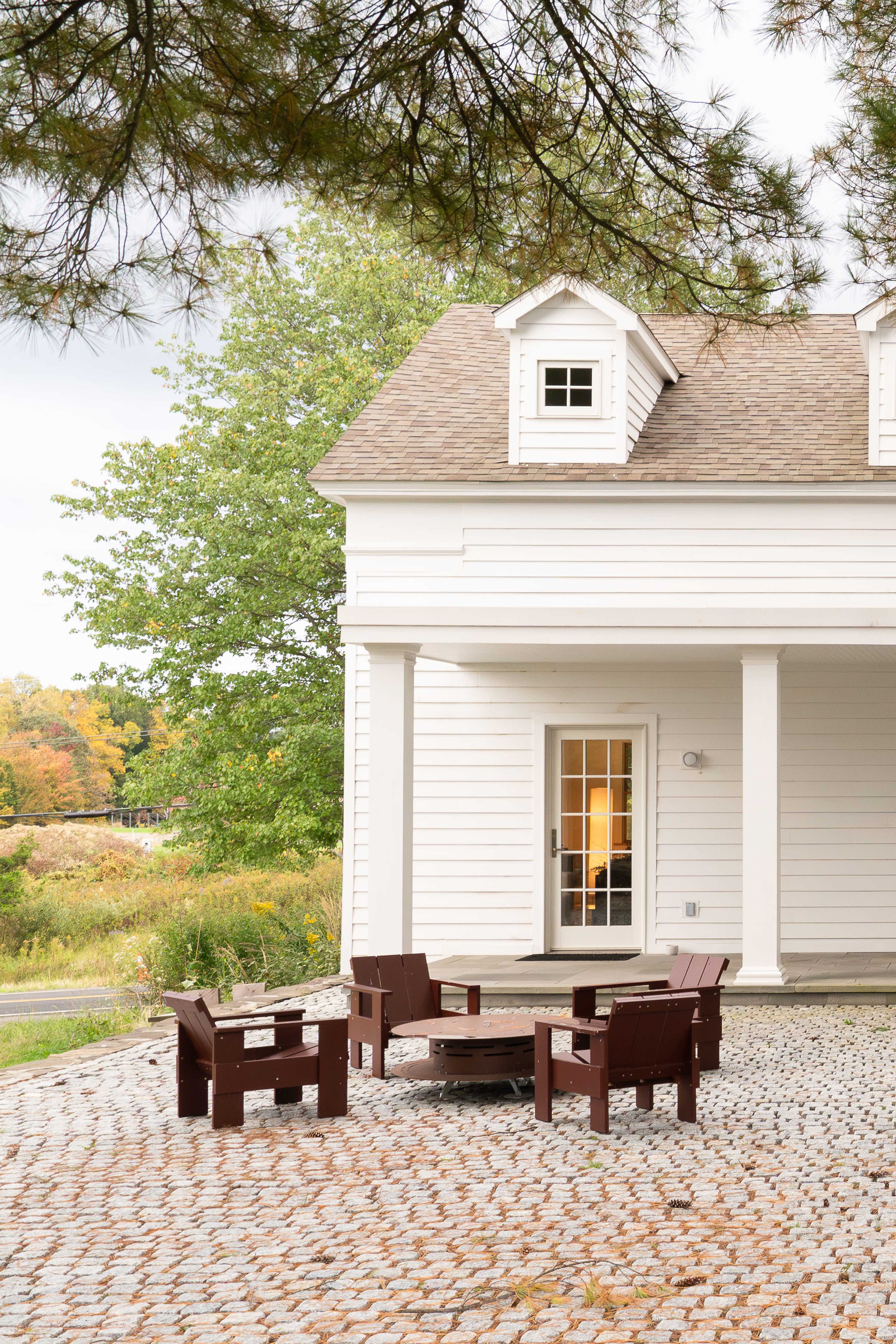 A seating area with brown wooden chairs surrounds a fire pit in front of a white house with a sloped roof.