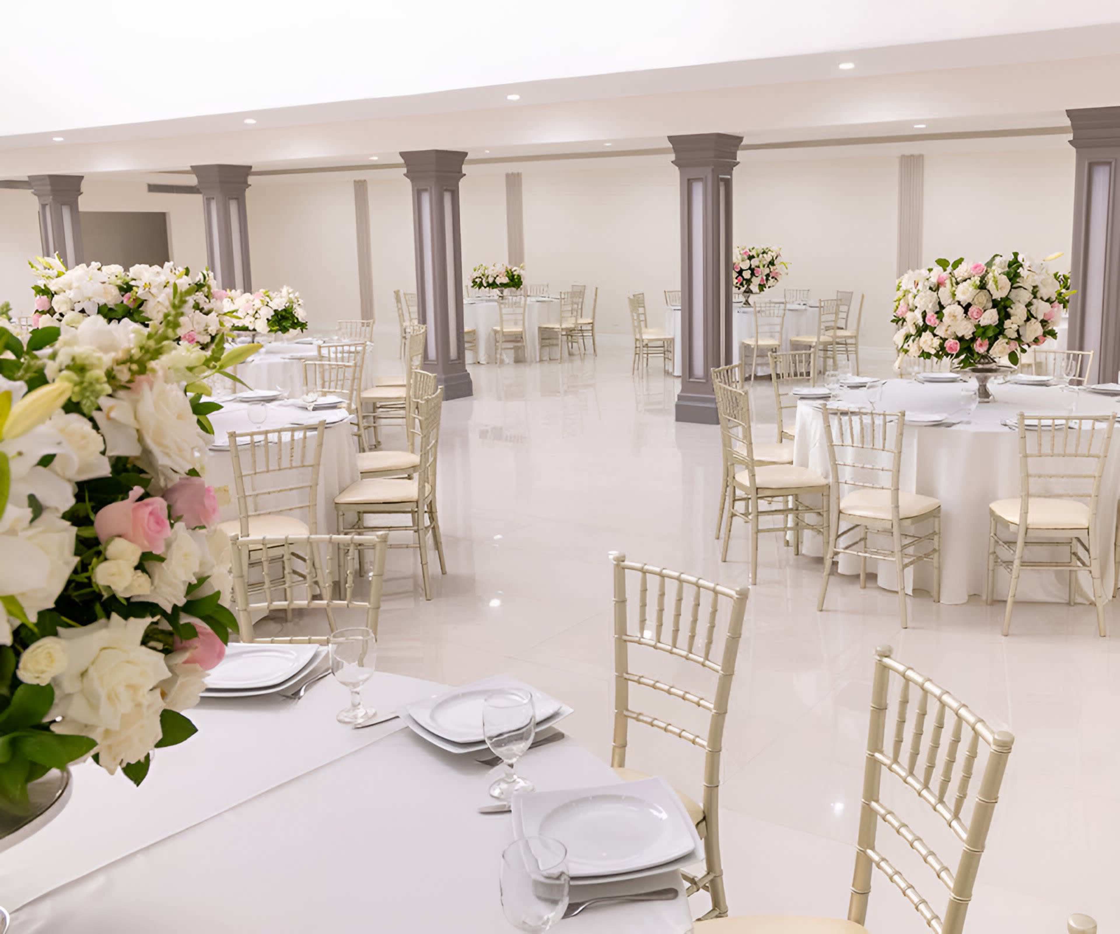 A spacious banquet hall is arranged with elegantly set round tables adorned with floral centerpieces and surrounded by gold-accented chairs.