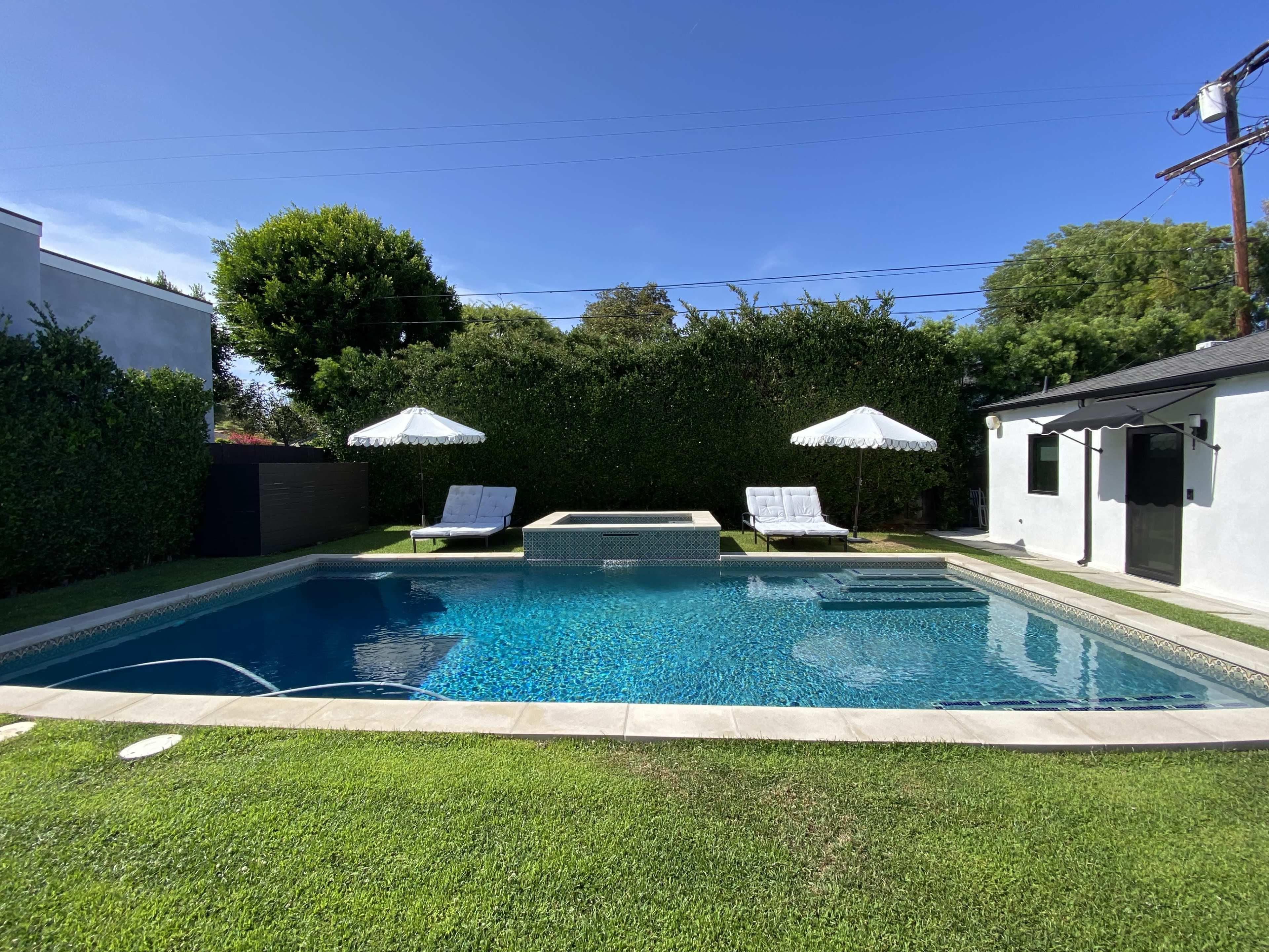 A backyard pool area with two sun loungers and a hot tub surrounded by greenery and a house in the background.