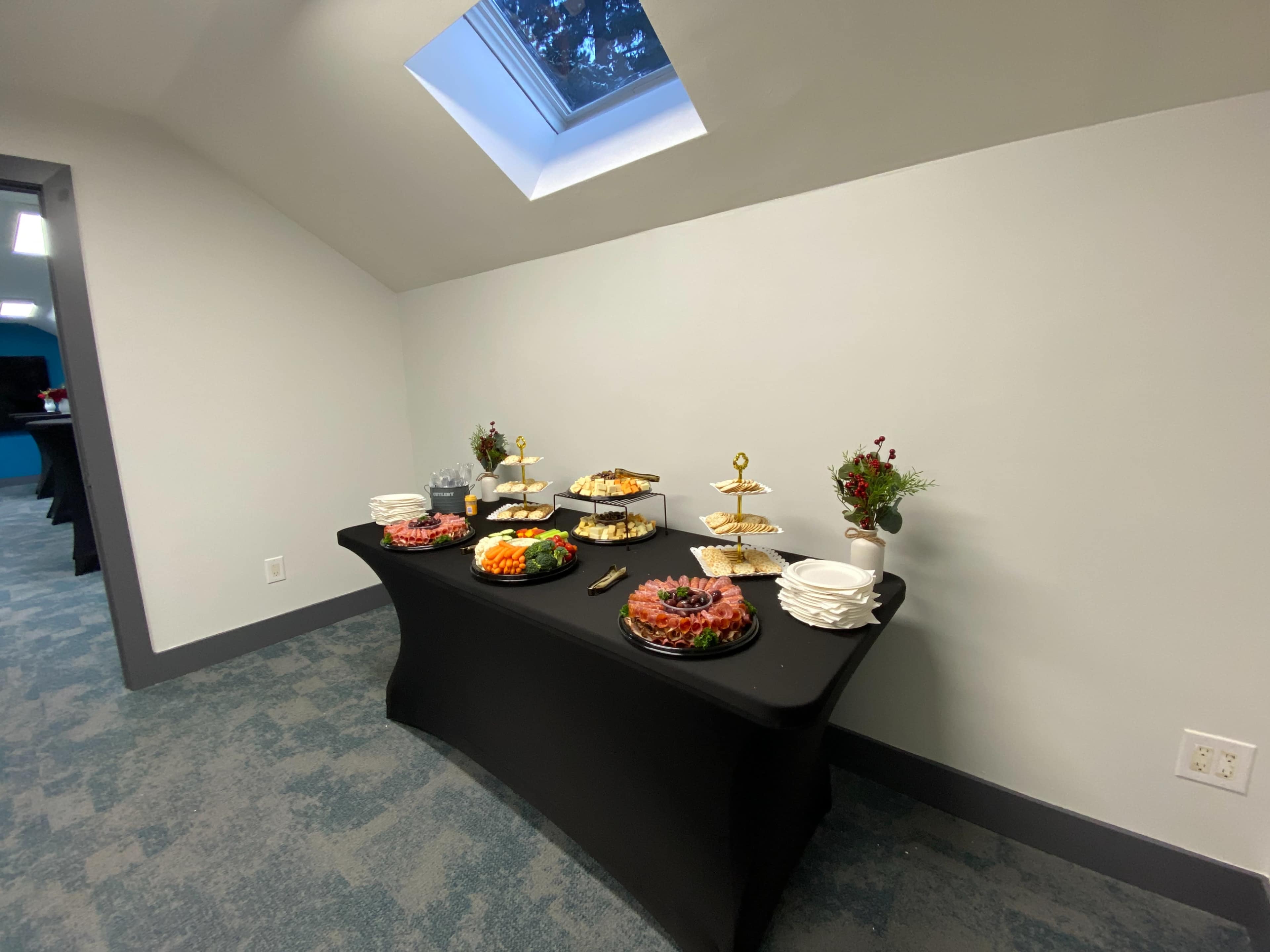 A black table is set with various food platters and snacks, surrounded by walls and a window in a room.