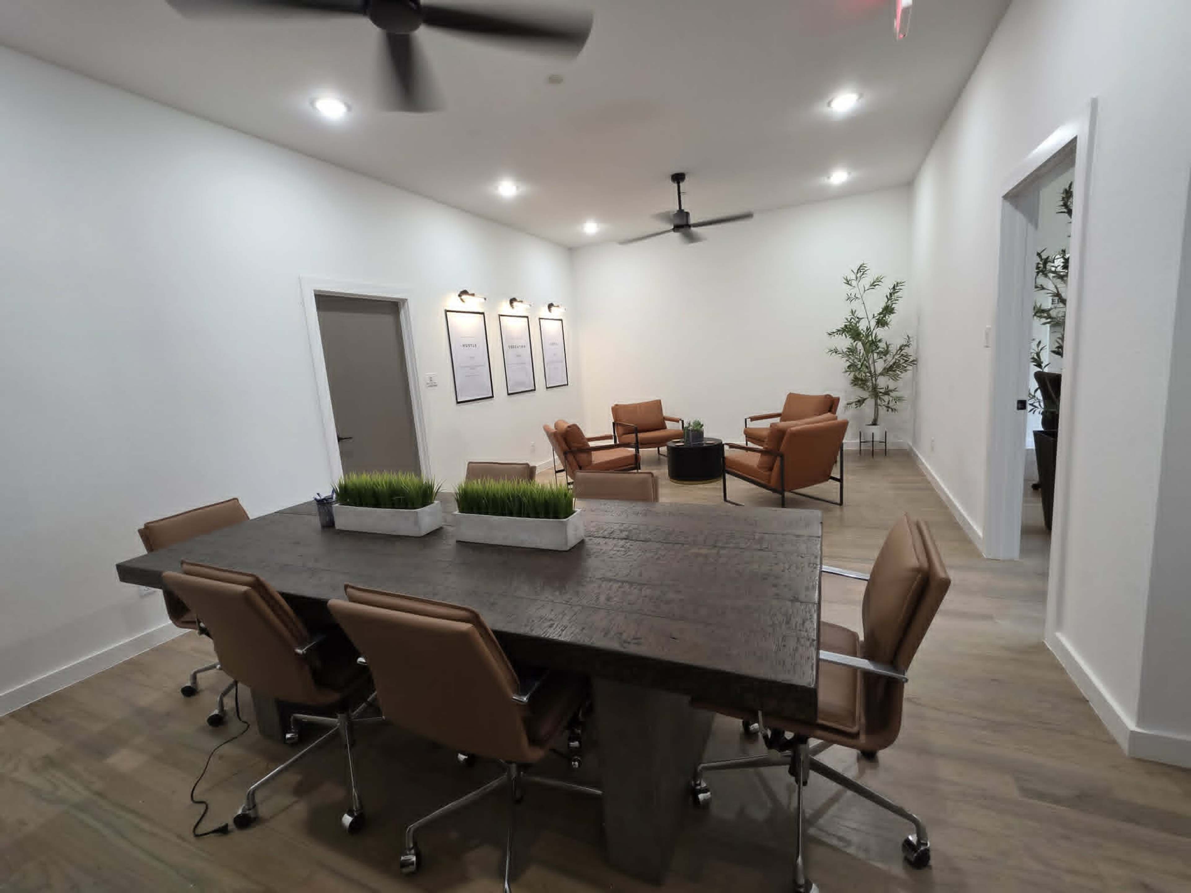 The image shows a modern conference room with a large wooden table surrounded by six rolling chairs, and two leather armchairs positioned nearby.