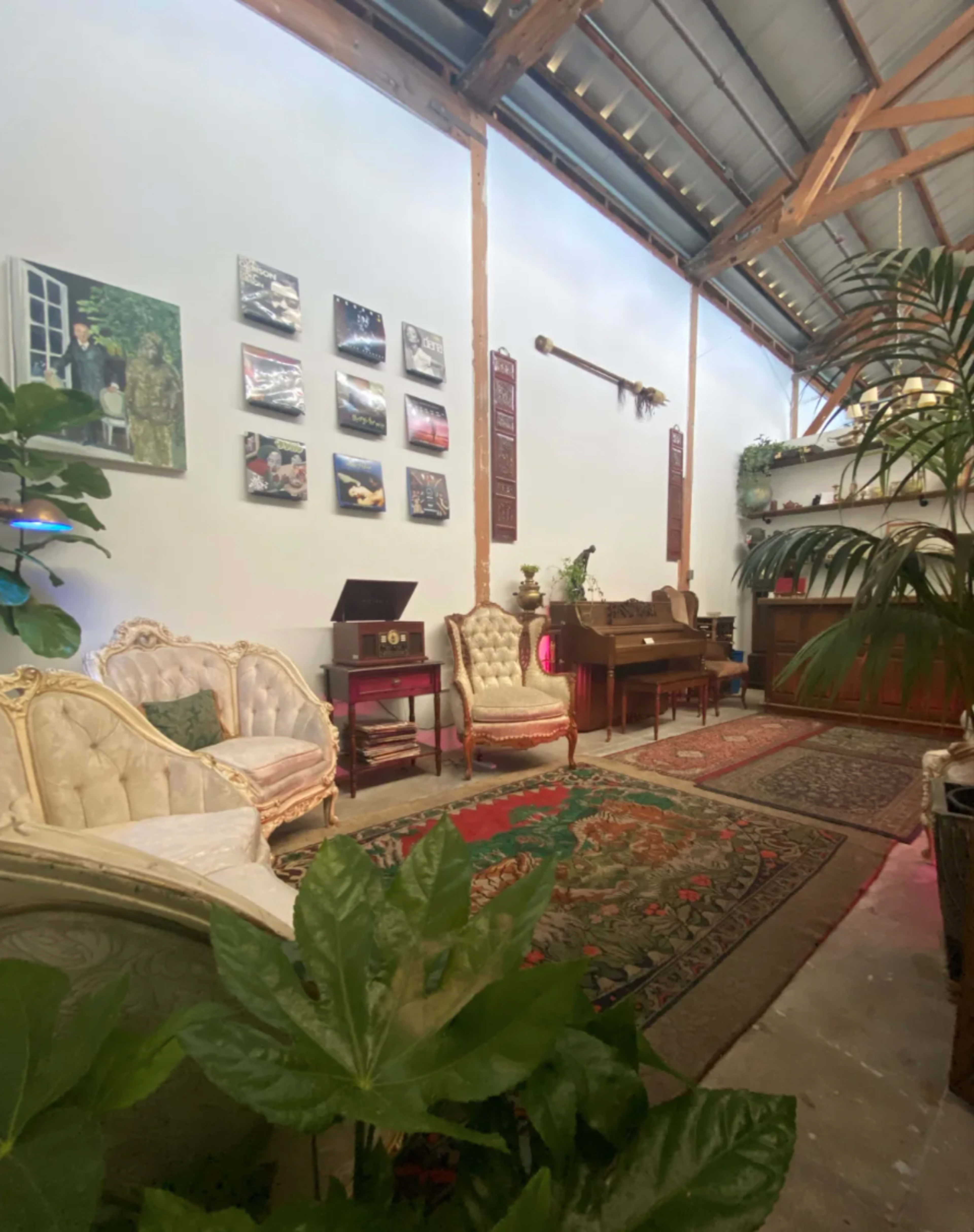 A spacious room with vintage-style furniture, including two ornate armchairs and a wooden piano, surrounded by plants and wall art.