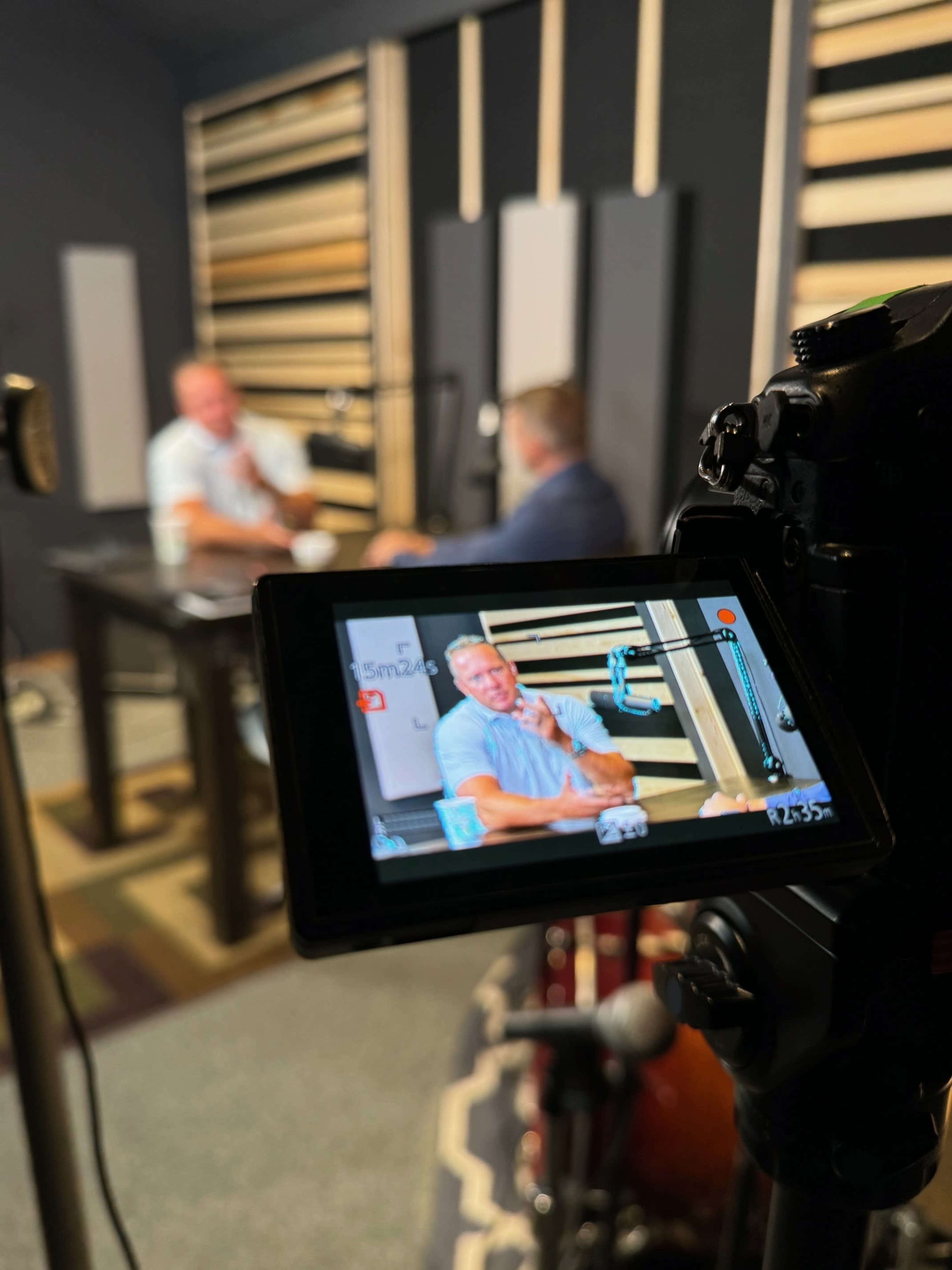 The image shows a camera viewfinder displaying two men sitting at a table in a podcast studio with soundproofing panels in the background.