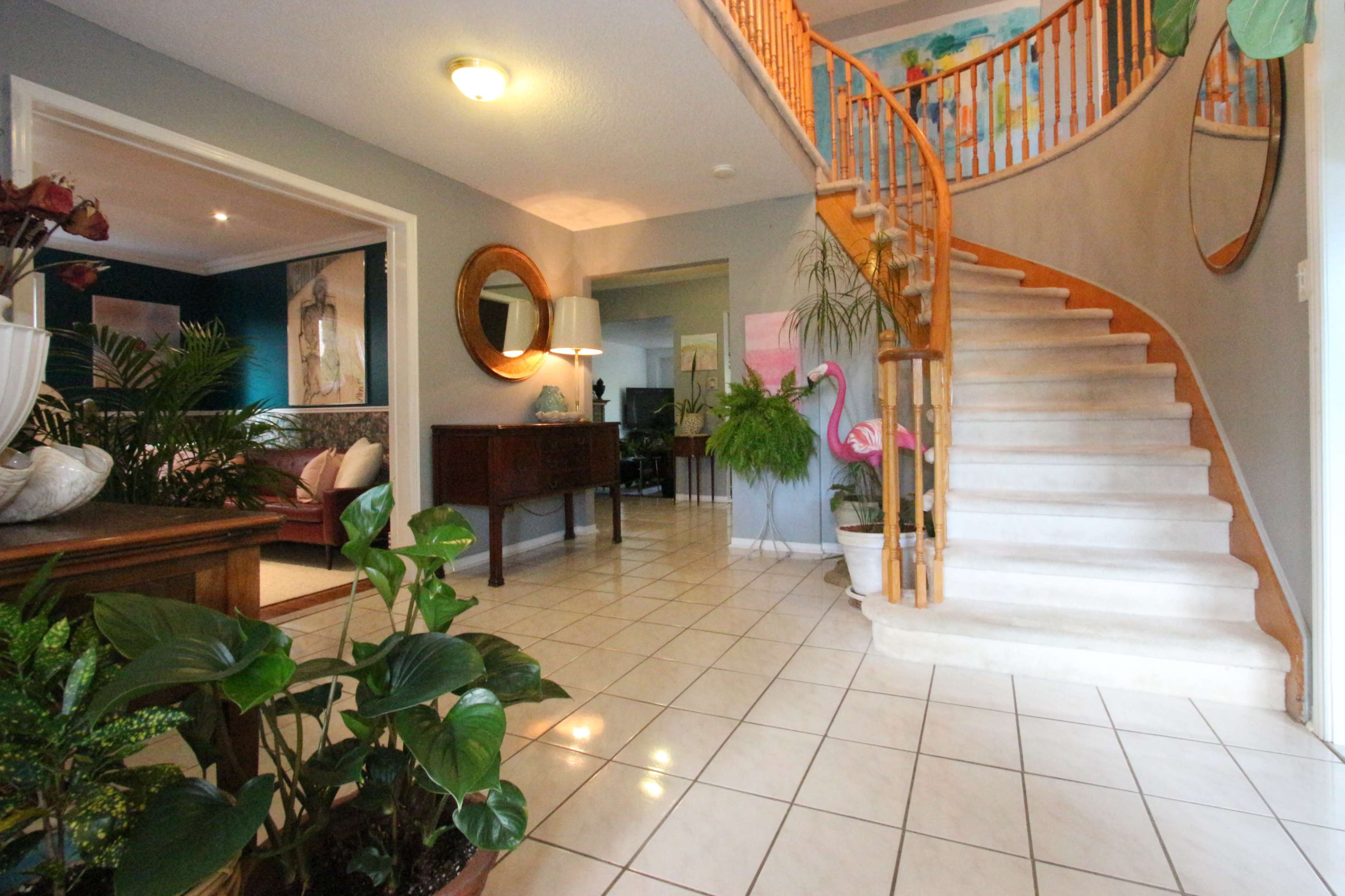 A well-lit entryway features a staircase, a wooden side table with plants, and a view into adjacent rooms.