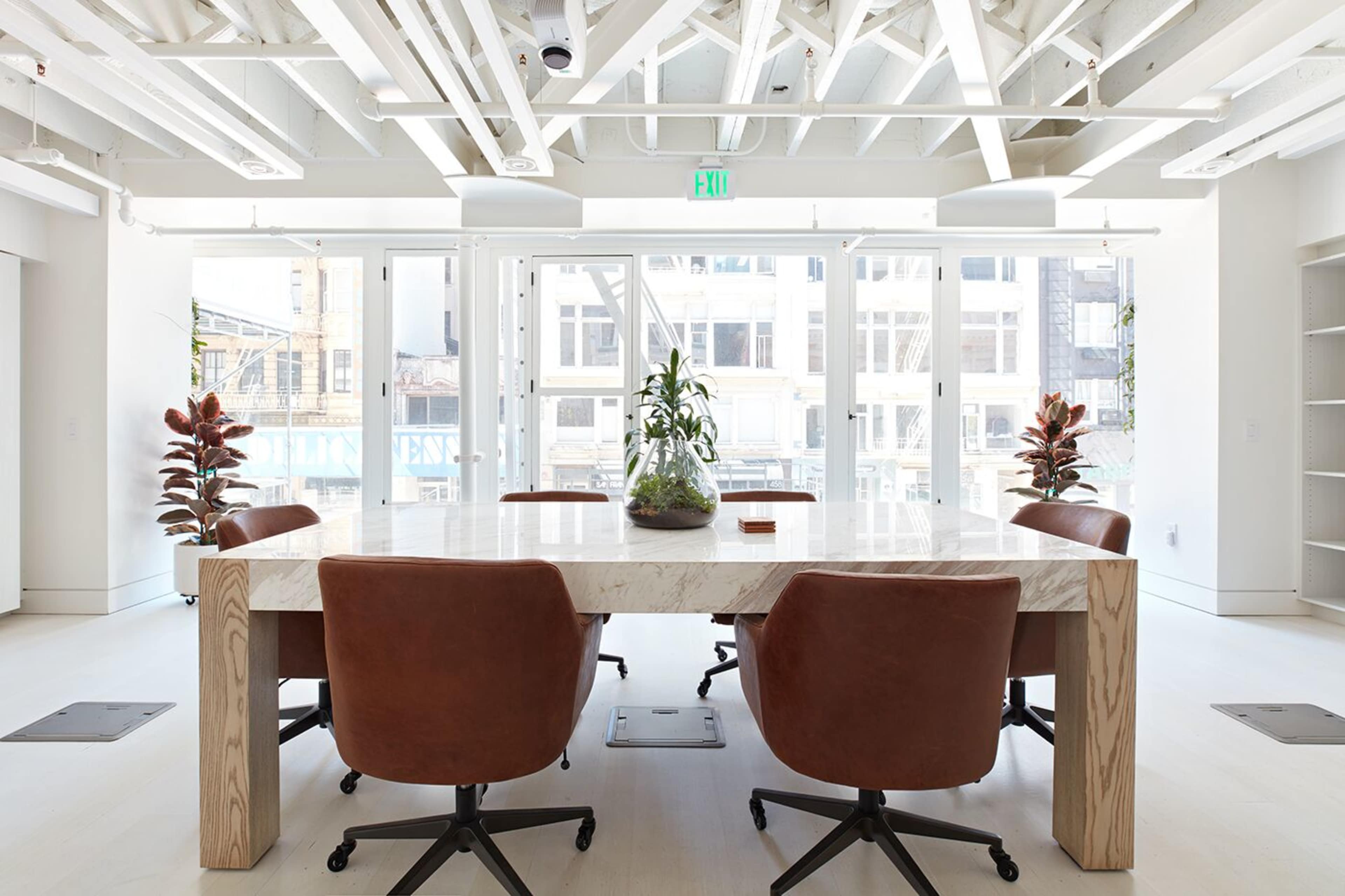 A spacious conference room features a large wooden table surrounded by brown leather chairs, with large windows allowing natural light and views of the city outside.
