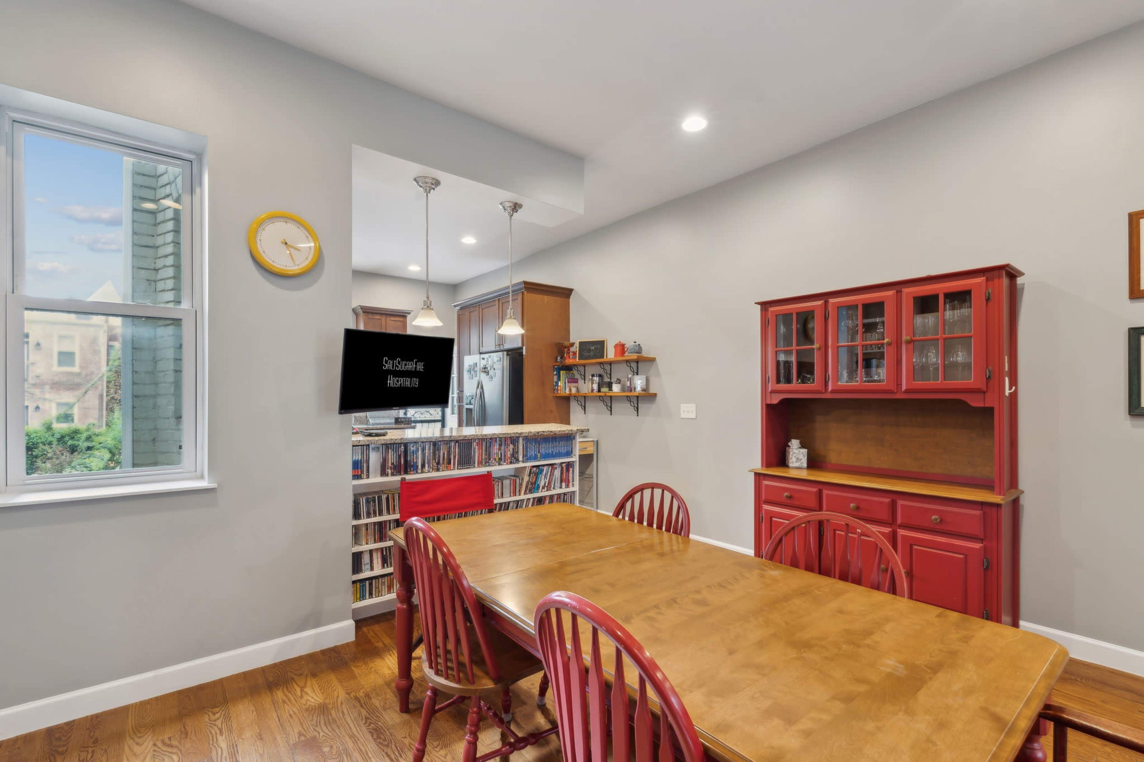 The image shows a modern dining area with a wooden table surrounded by red chairs, a red cabinet, and a television mounted on the wall.