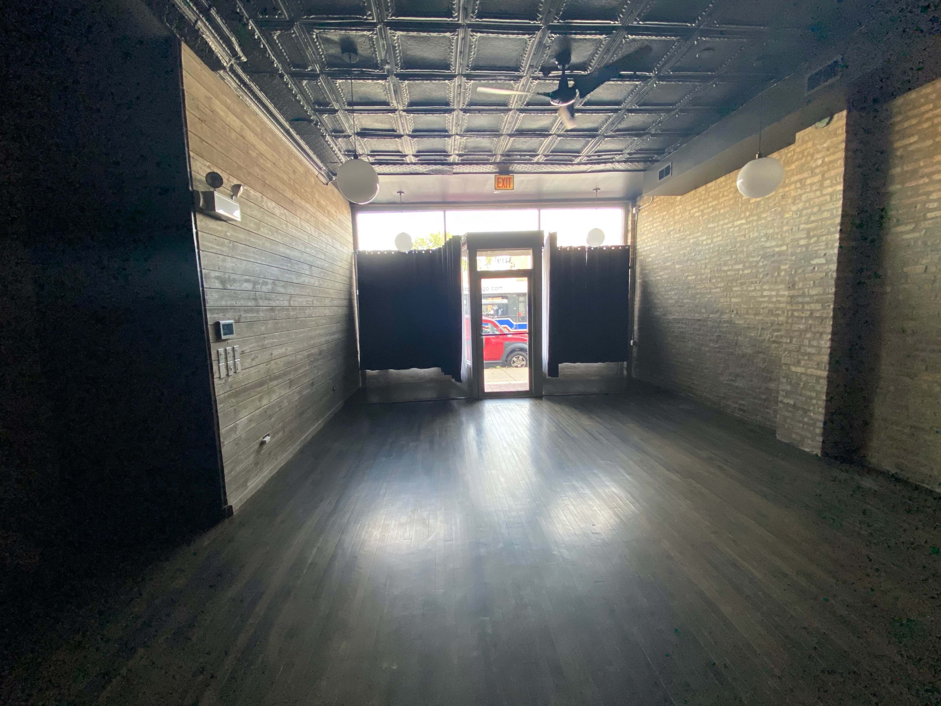 An empty interior space with wooden flooring, exposed brick walls, and a set of double doors leading outside.