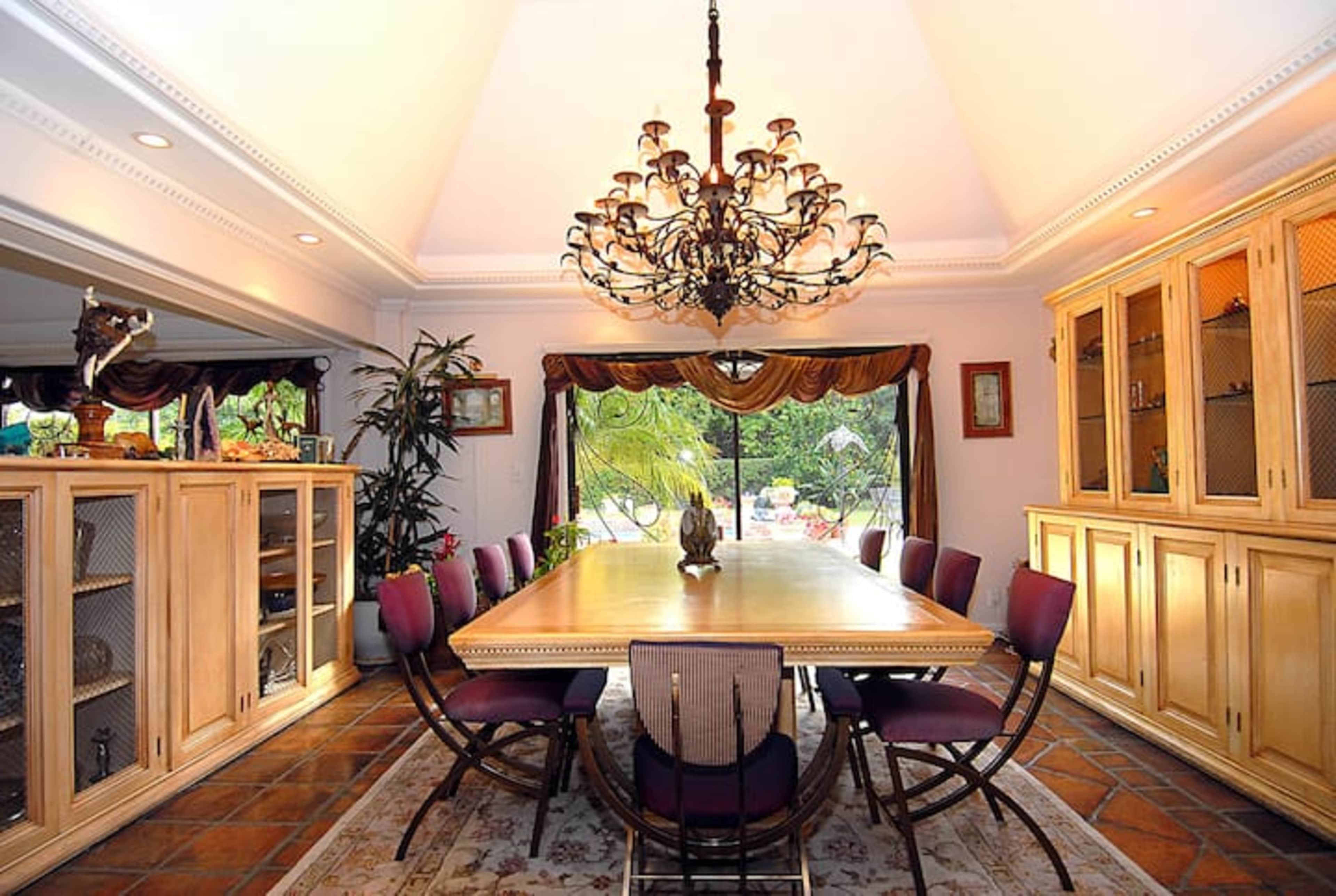 A spacious dining room features a large wooden table surrounded by chairs, with a decorative chandelier hanging above and a window offering a view of a garden.